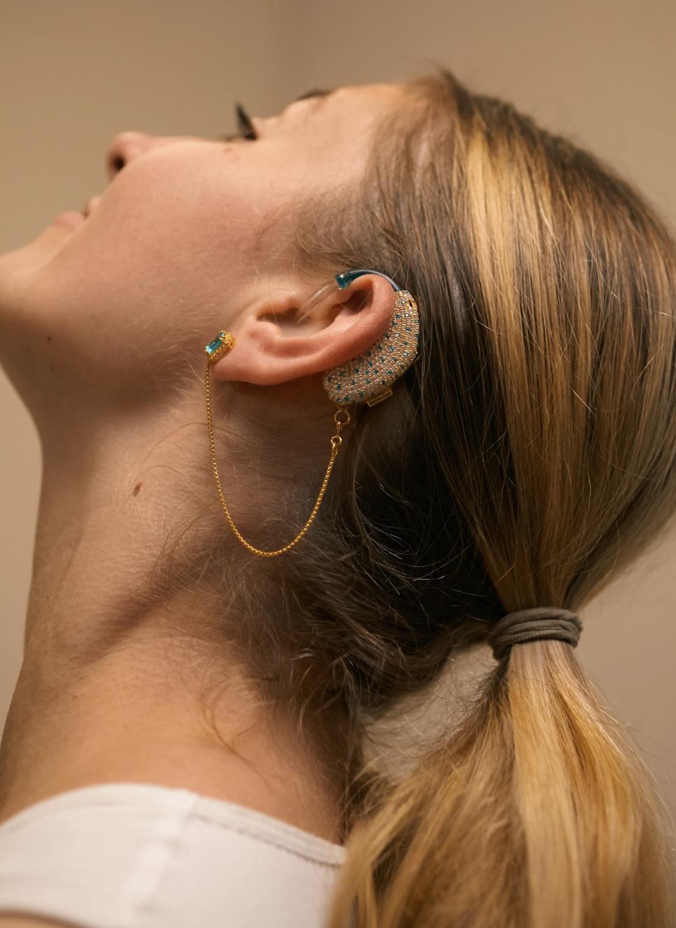 Side profile of a woman wearing a crystal-covered gold AUZI hearing-aid cover connected by a fine chain to a blue gemstone earring