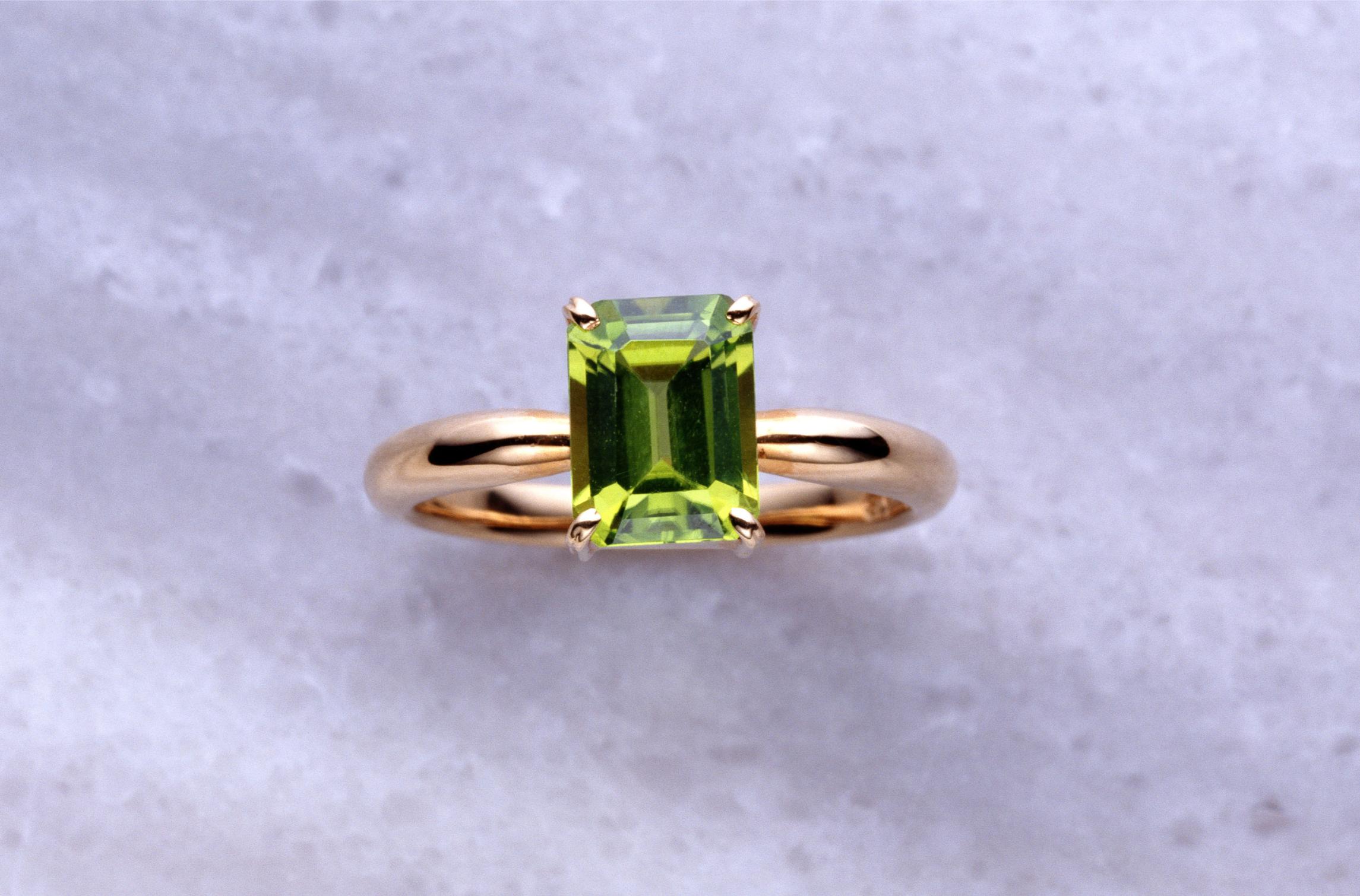 ellow-gold solitaire ring featuring a rectangular emerald-cut green peridot held in four prongs, photographed against a pale textured background.