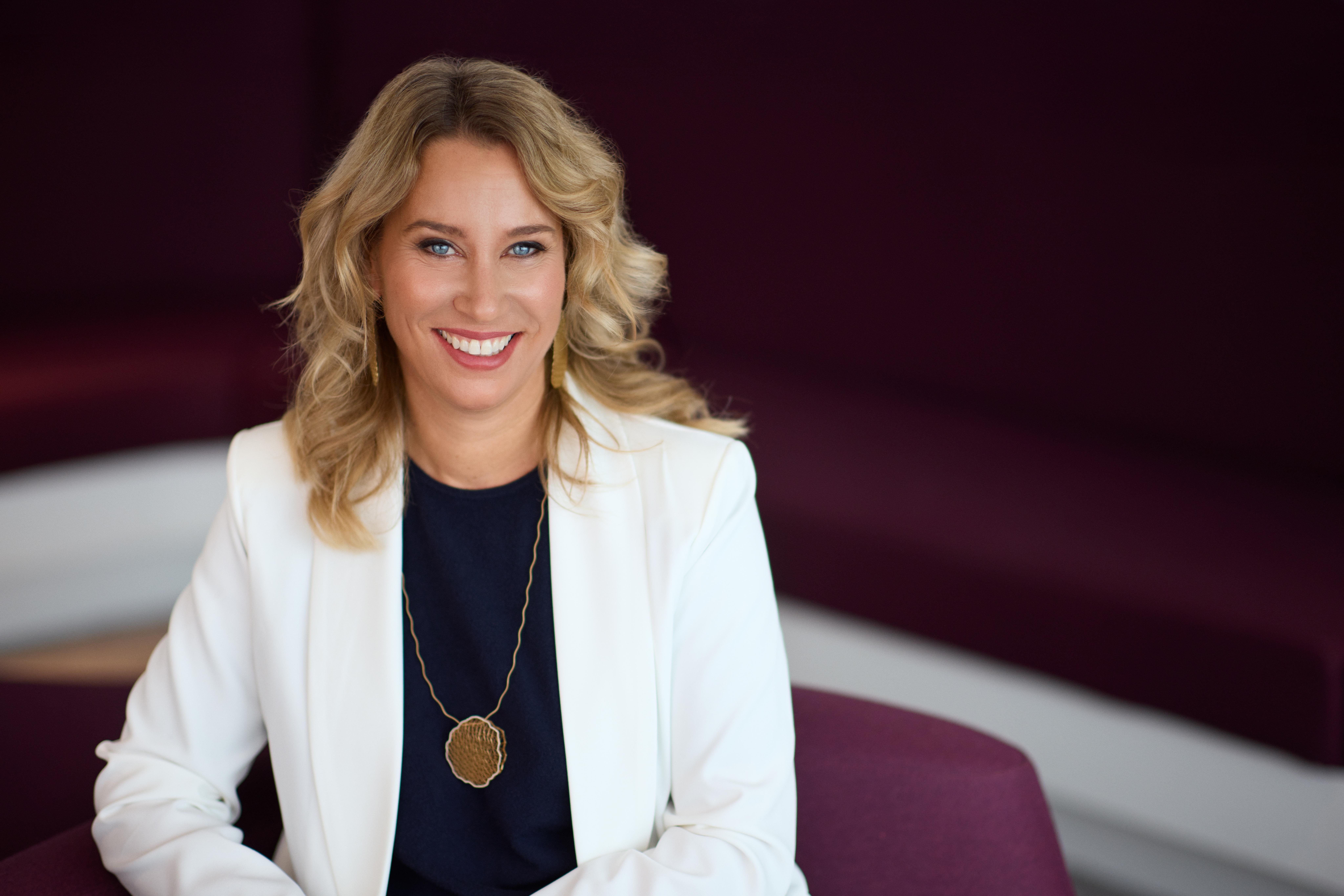 Portrait of INHORGENTA Exhibition Director Stefanie Mändlein wearing a white blazer, dark top and long gold pendant necklace.