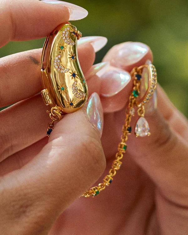 Close-up of bespoke gold AUZI hearing-aid covers decorated with diamond crescents and blue and green stars, joined by a fine gemstone-set chain