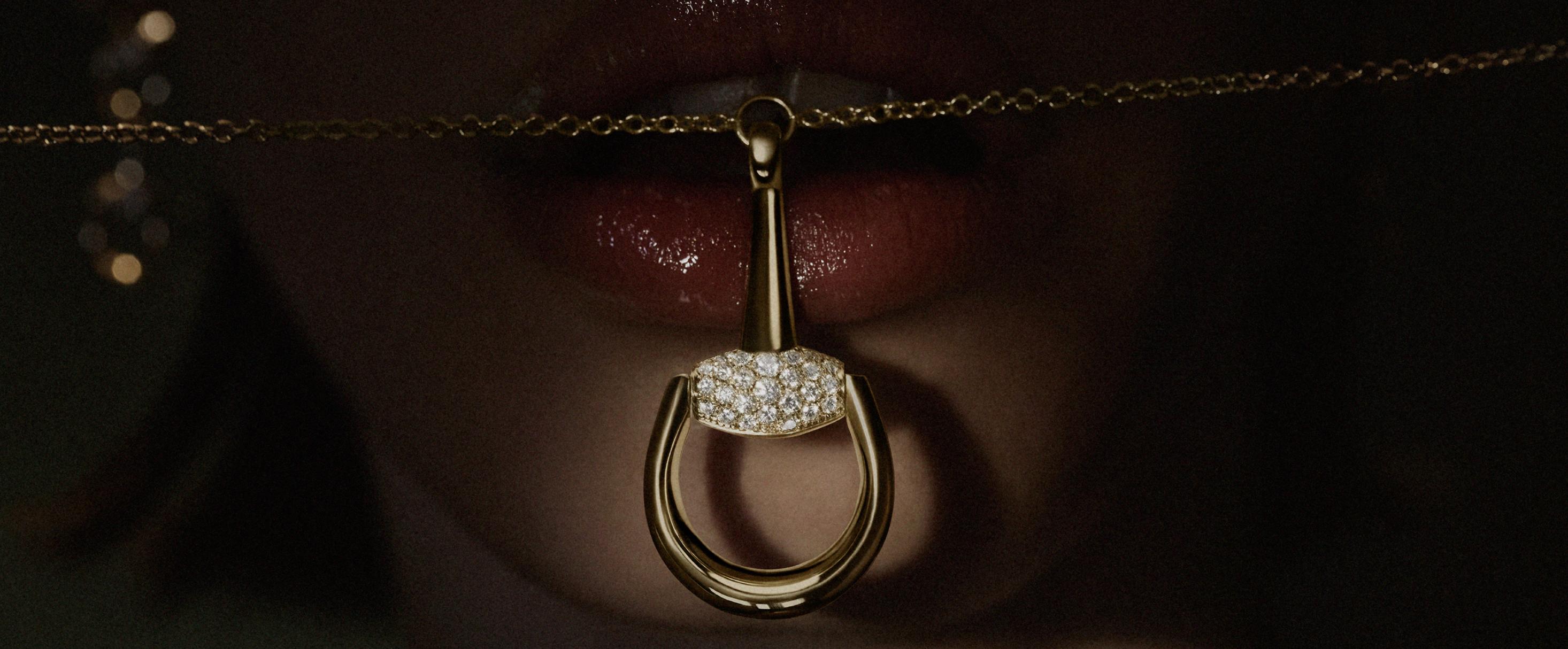 Close-up of a diamond-set yellow-gold Gucci Horsebit pendant necklace held between a model’s lips against a dark background