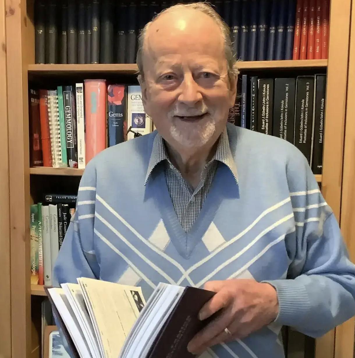 Portrait of renowned gemologist Alan Hodgkinson holding an open gemology book in front of a library of reference books. The Alan Hodgkinson Medal honours his lasting legacy of education, mentorship and outstanding contribution to the gemological profession.
