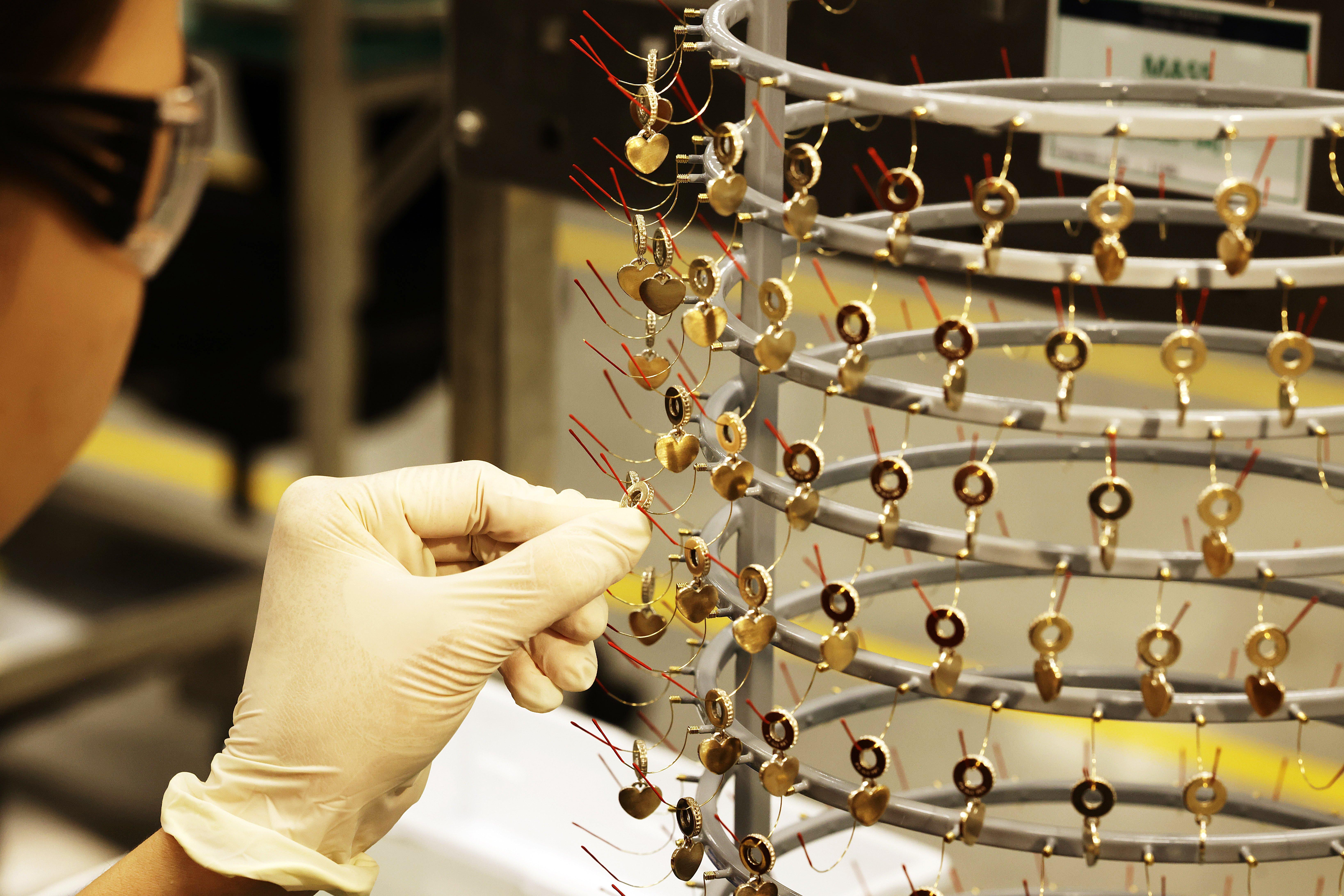 A gloved craftsperson examines rows of gold-coloured heart charms suspended from a circular production rack.