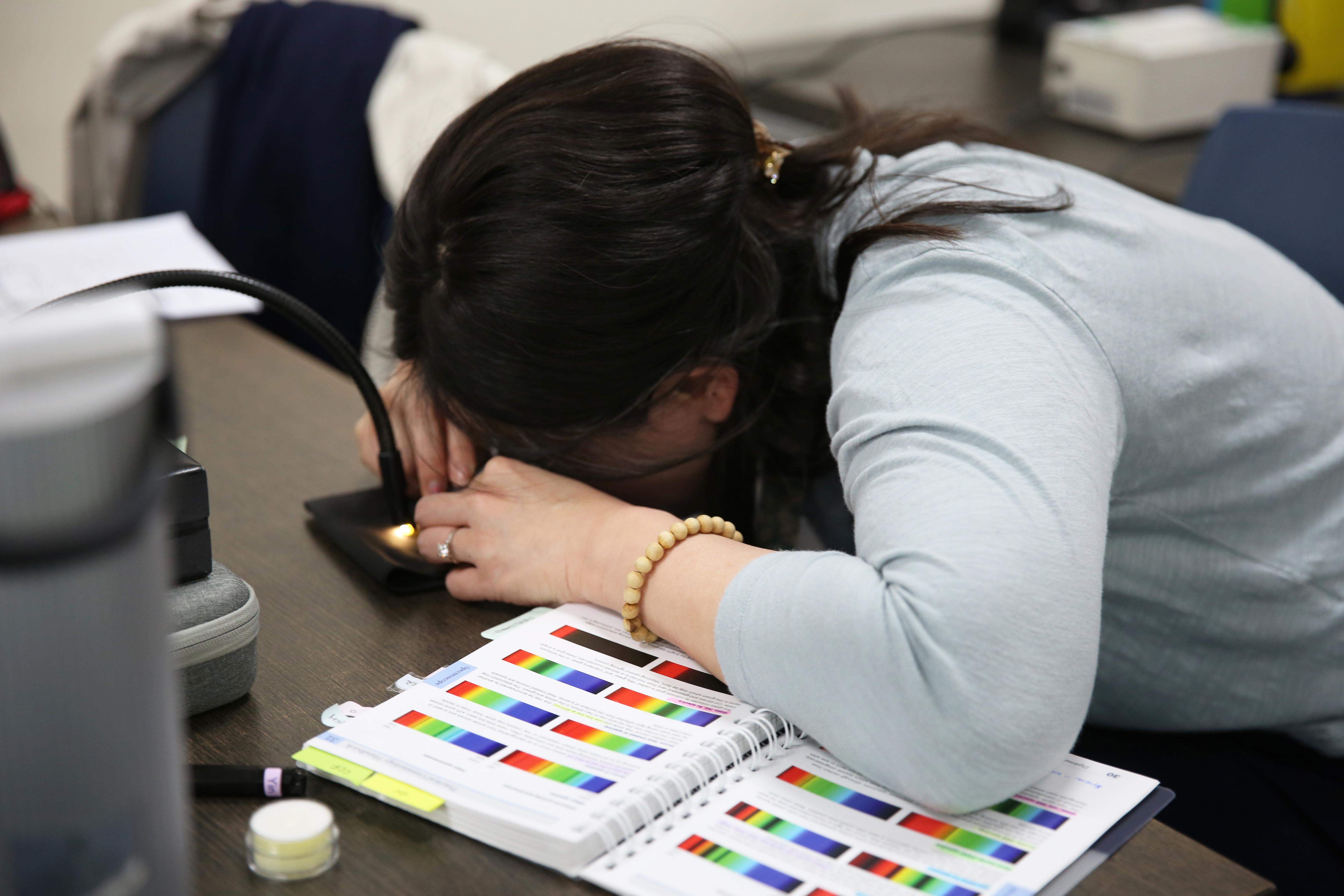 Gem-A student examining a gemstone under a light beside a gemmology reference book in a London classroom.