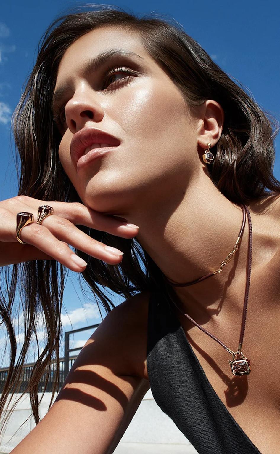 Model wearing Stephen Webster Full Bleed rings, a drop earring and a gemstone padlock pendant on a leather cord against a blue sky.