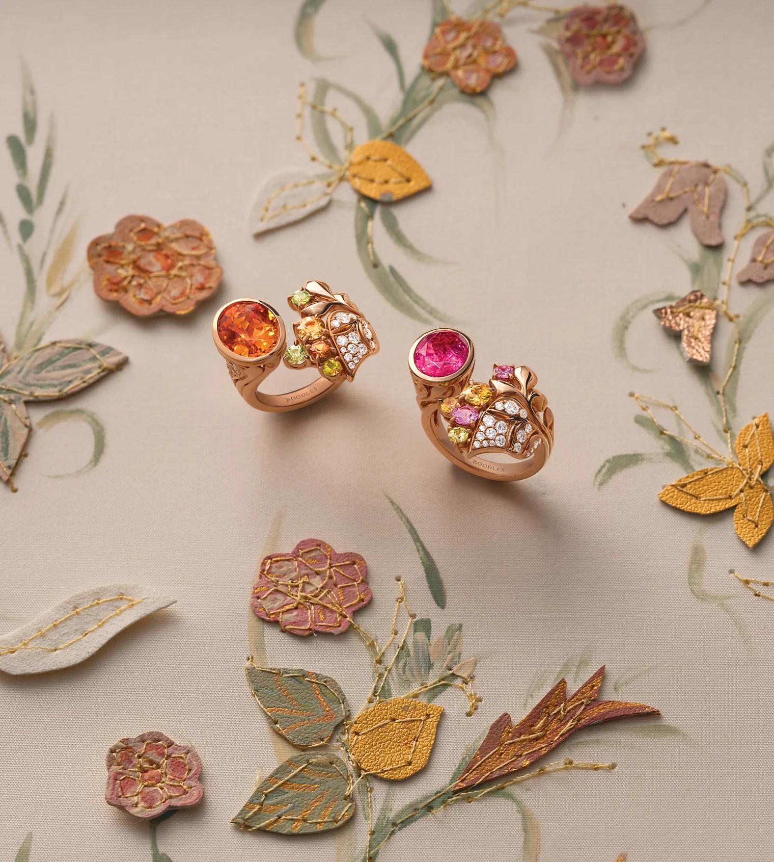 Two Silk Mantua rings from Boodles’ Palace Collection featuring pink tourmalines, mandarin garnets, coloured sapphires and diamonds set in rose gold, displayed against a backdrop inspired by the floral motifs of a historic Spitalfields silk mantua.
