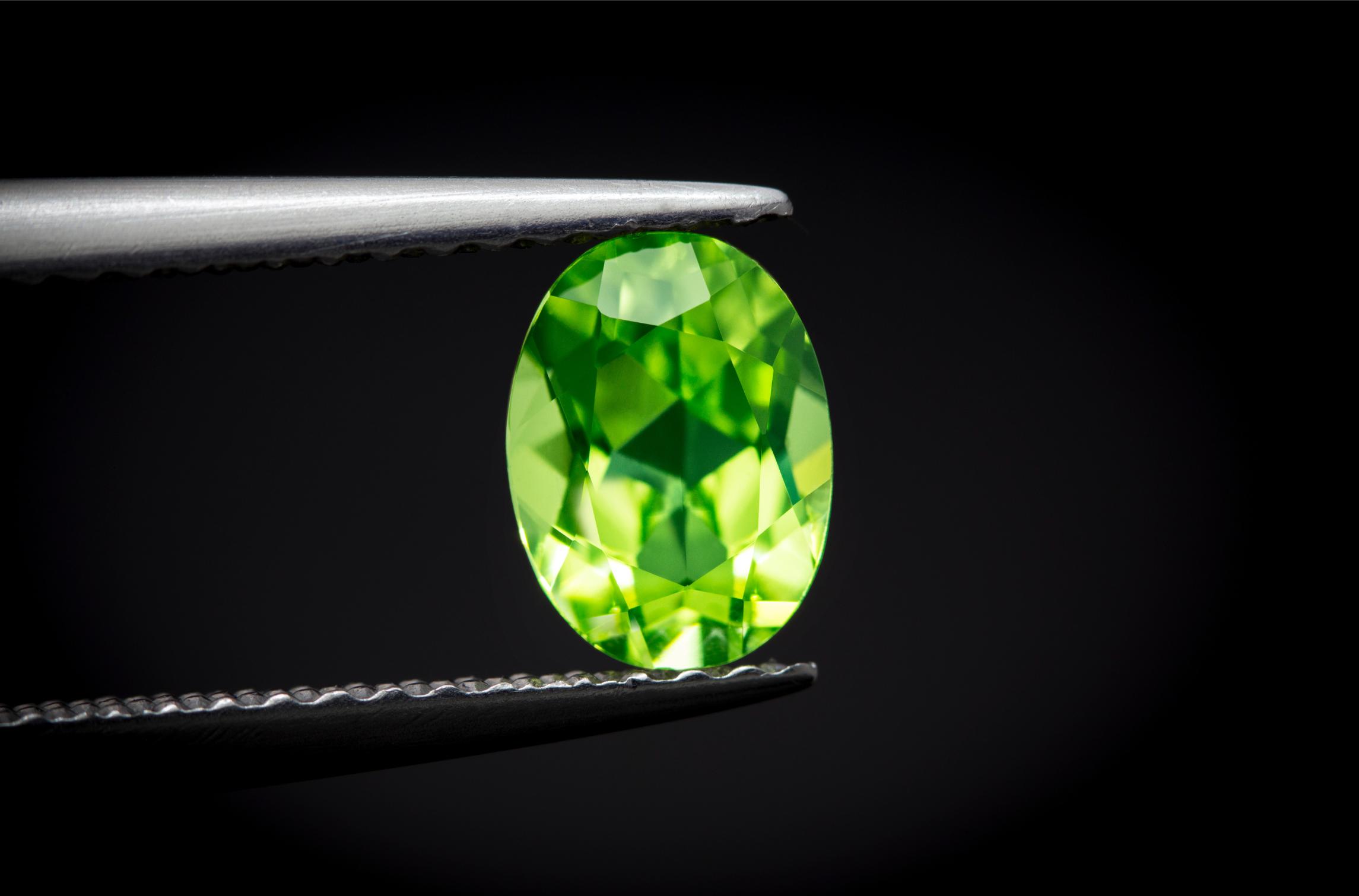 Close-up of a vivid green oval-cut peridot held between metal tweezers against a black background, highlighting its brilliant faceting and yellow-green flashes.