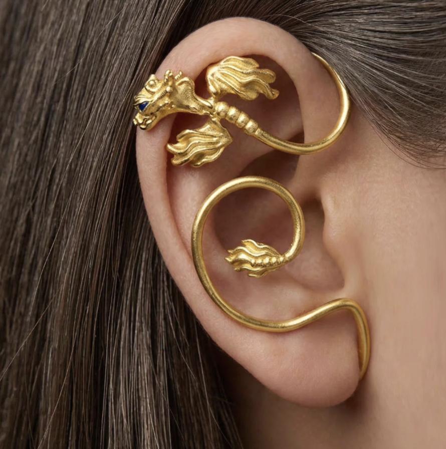 Close-up of a gold-vermeil dragon ear wrap curling around a model’s ear, with sculpted wings and small blue gemstone eyes.
