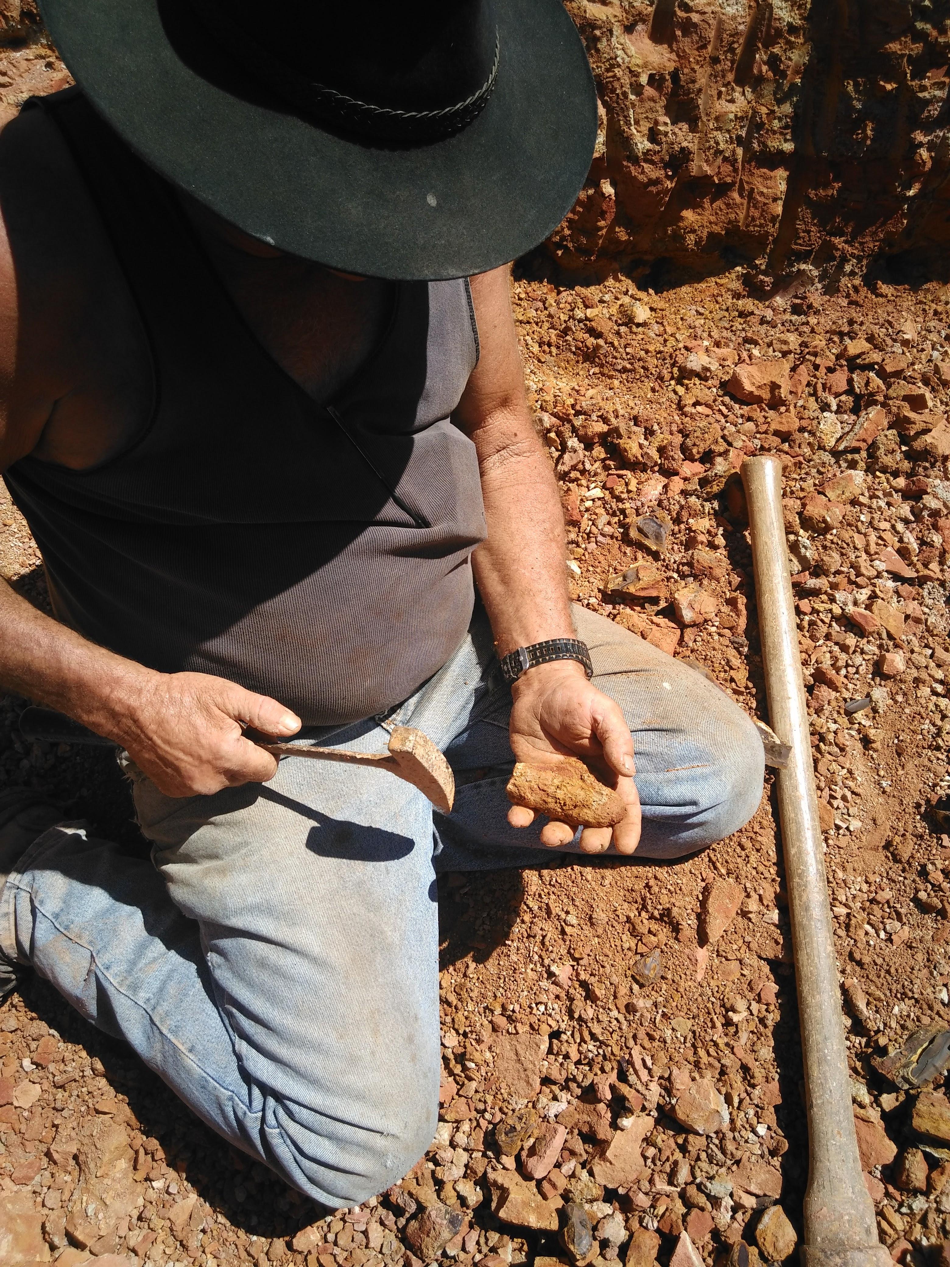 An opal miner kneels on red earth holding a piece of rough ironstone and a hand tool at a mine in western Queensland