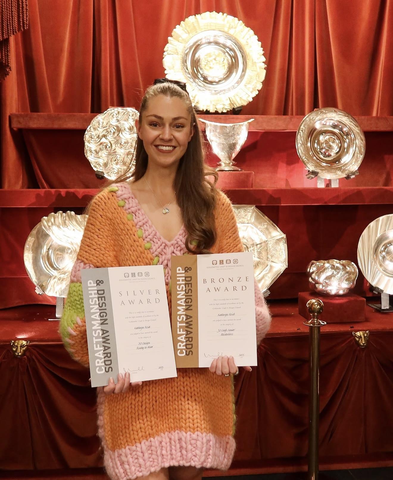 Jewellery designer Kathryn Reid smiling and holding silver and bronze Craftsmanship and Design Award certificates in Goldsmiths’ Hall.