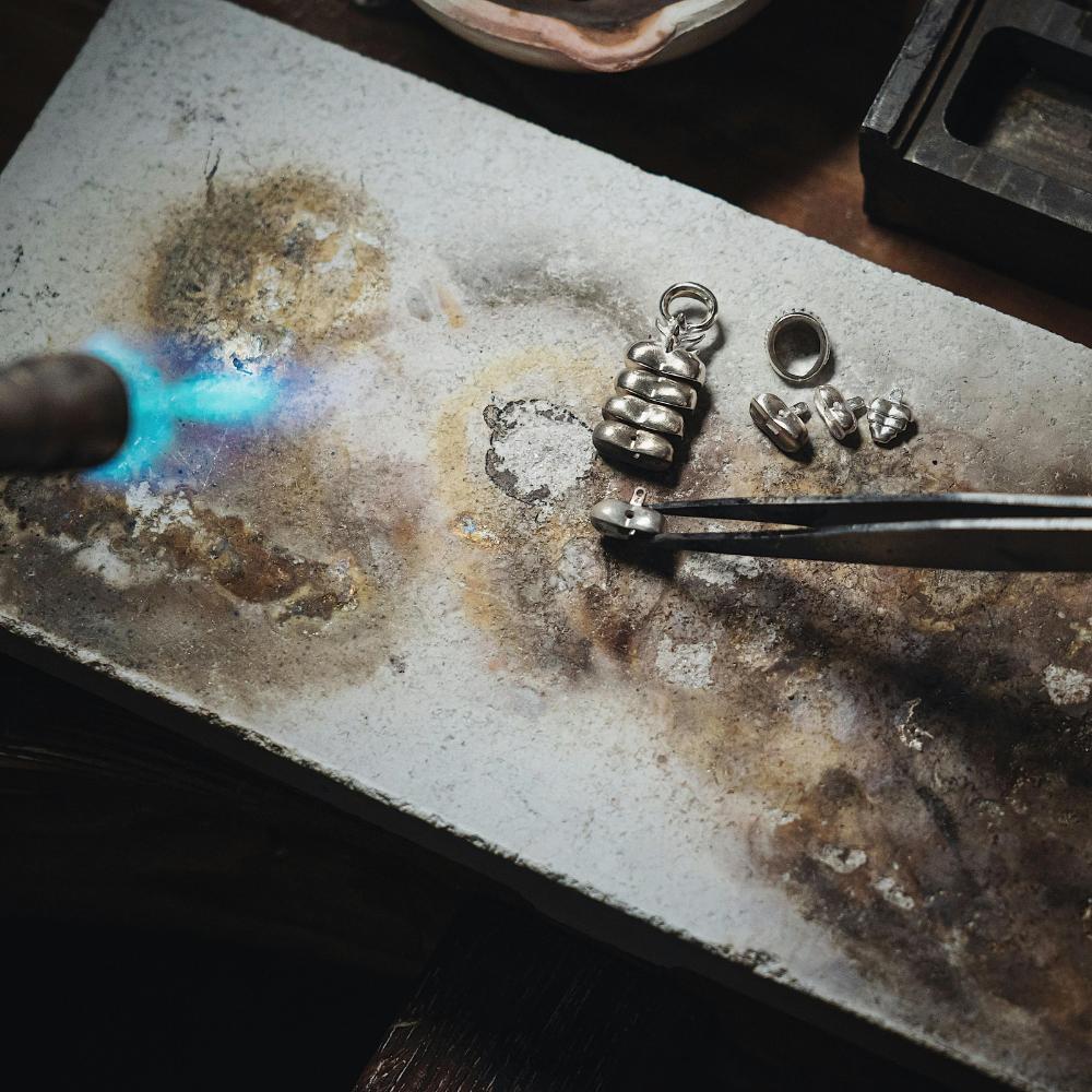A blue torch flame heats small silver jewellery components held with tweezers on a scorched soldering block