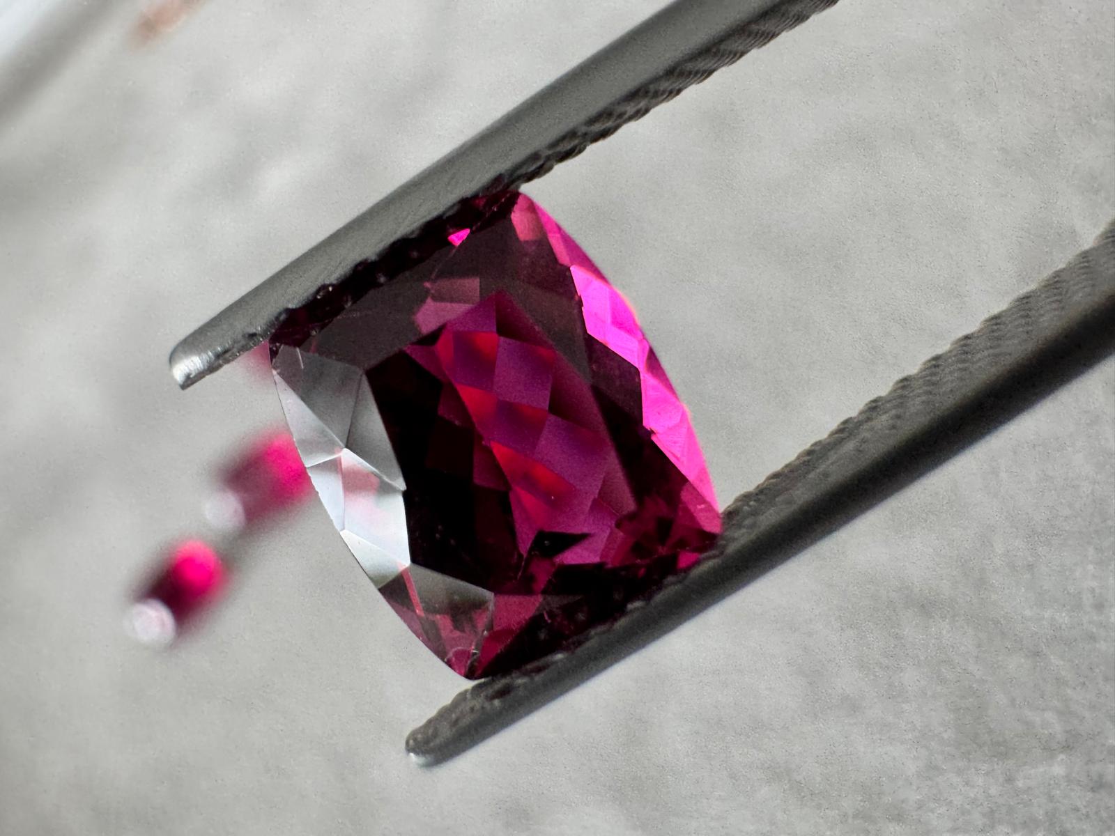 A vibrant pink gemstone held by tweezers, showcasing its cushion cut and vivid clarity against a soft grey background.