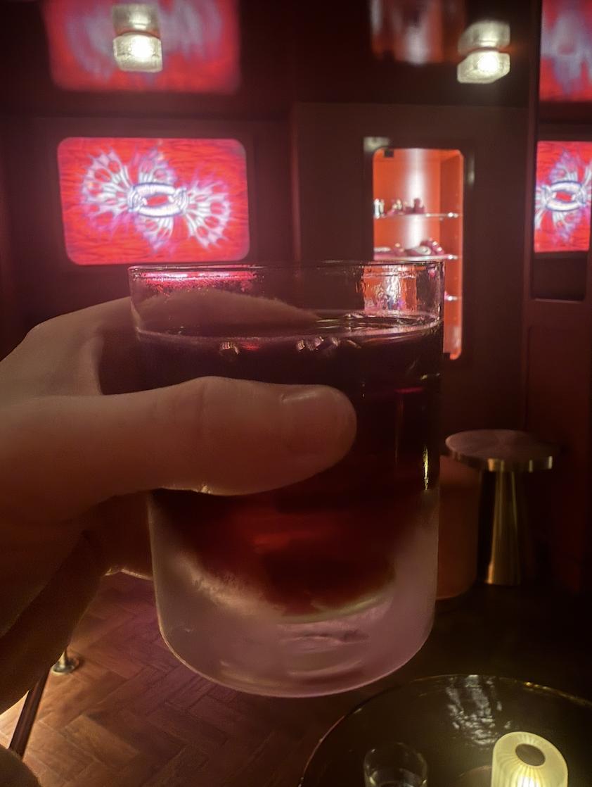 Hand holding a Ruby Negroni over ice inside Stephen Webster’s red-lit No Regrets Lounge in London