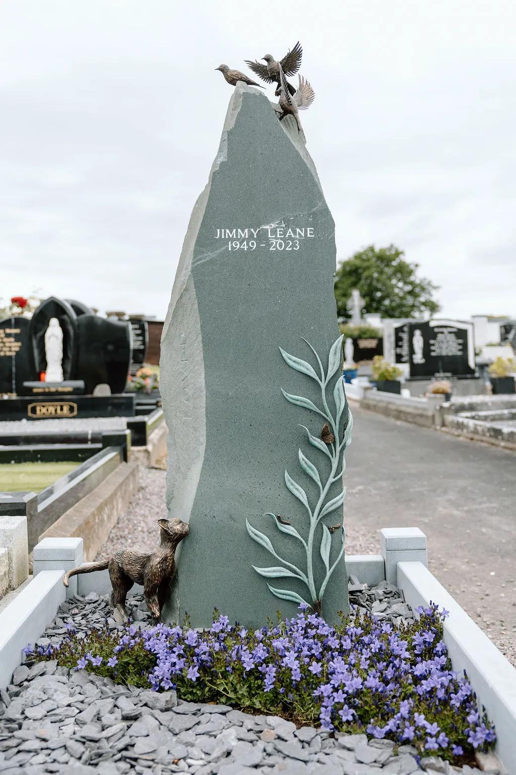 Tall grey gravestone dedicated to Jimmy Leane, decorated with sculpted birds, leafy branches, insects and a bronze cat, with purple flowers at its base