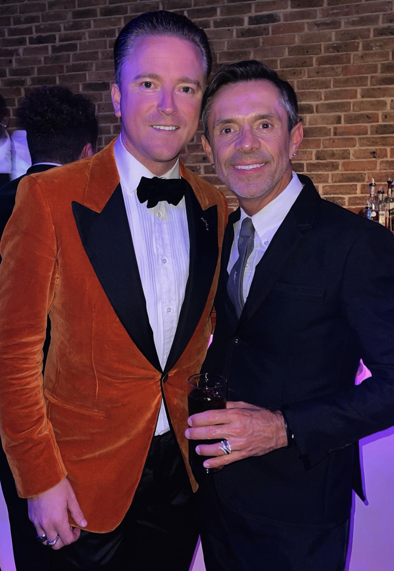 Andrew Martyniuk in an orange velvet dinner jacket standing beside Shaun Leane in a dark suit at an evening event