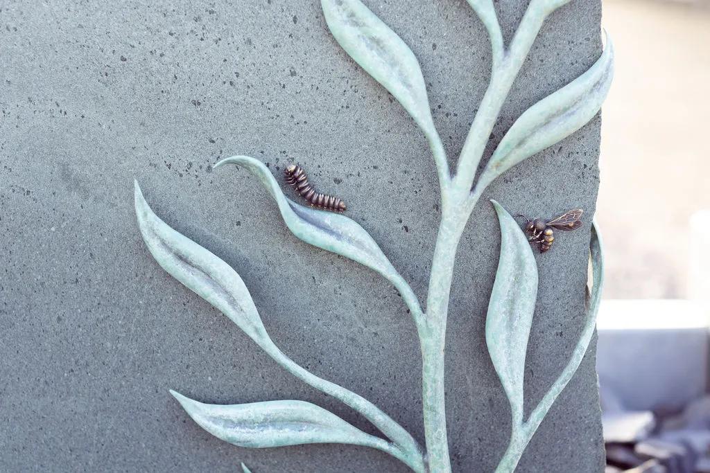 Close-up of patinated metal leaves, a caterpillar and a bee set against the grey stone memorial created by Shaun Leane for his father
