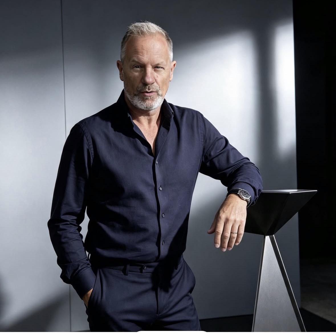 Portrait of Paul Henderson wearing a dark navy shirt and leaning against a geometric display stand in a modern interior