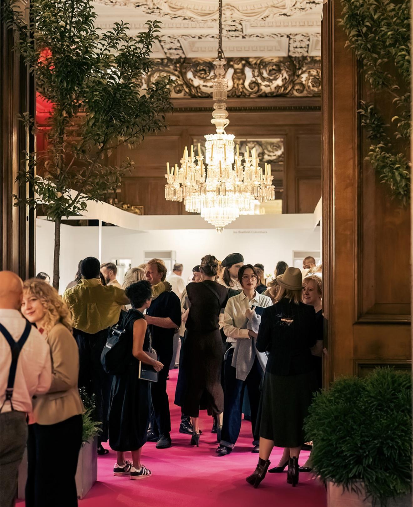 Visitors browsing Goldsmiths’ Fair inside the ornate Goldsmiths’ Hall, with a bright chandelier above the exhibition stands and a pink carpet below