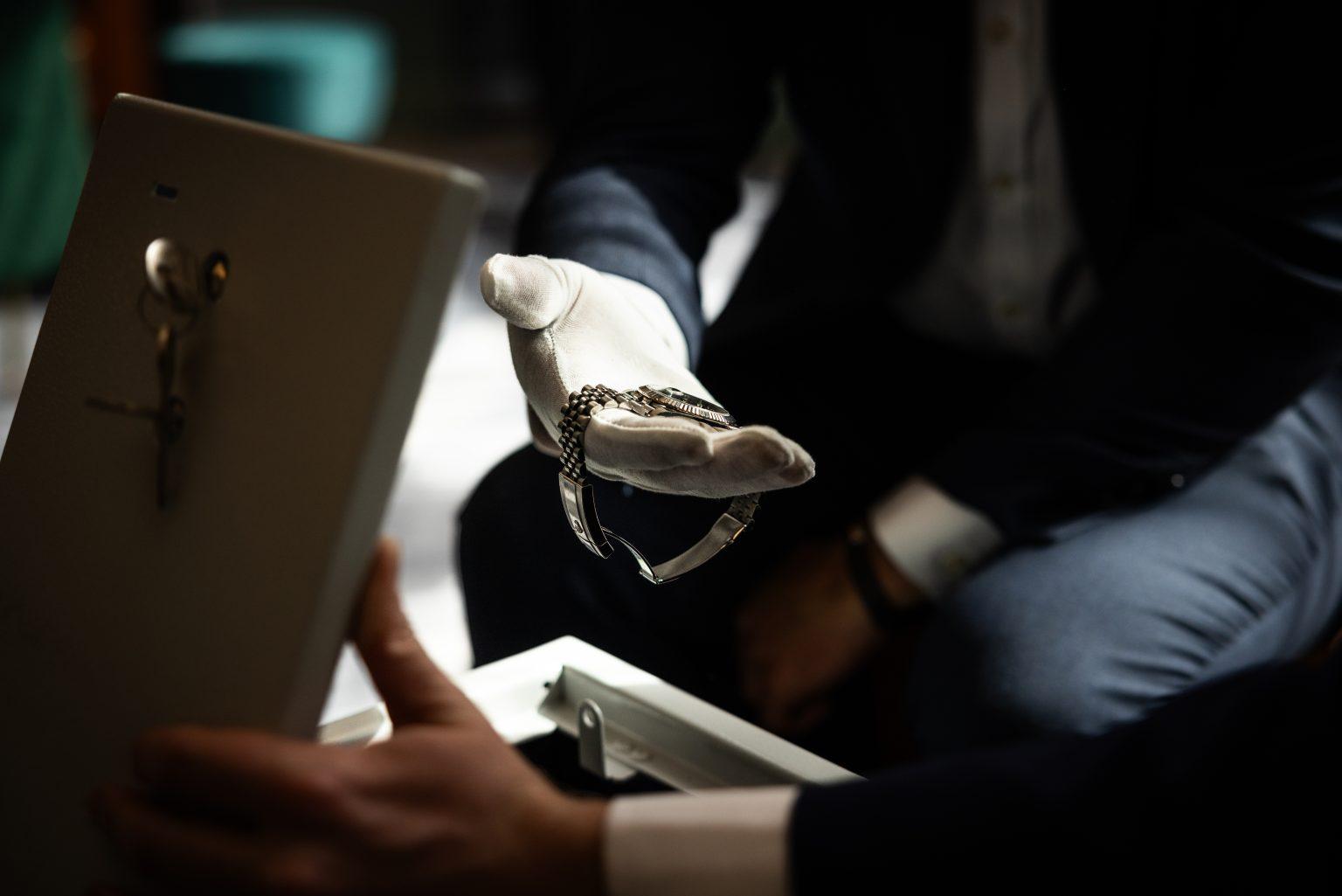 A gloved hand presenting a luxury watch beside an open secure storage case, illustrating UltraVault&rsquo;s handling and access service.