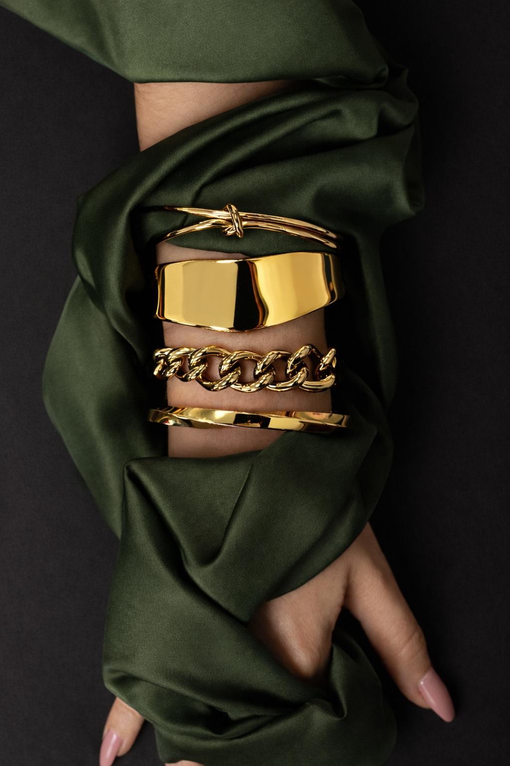 Model wearing four gold toned Ava & Sky bangles, including polished, chain and knot designs, over dark green fabric.