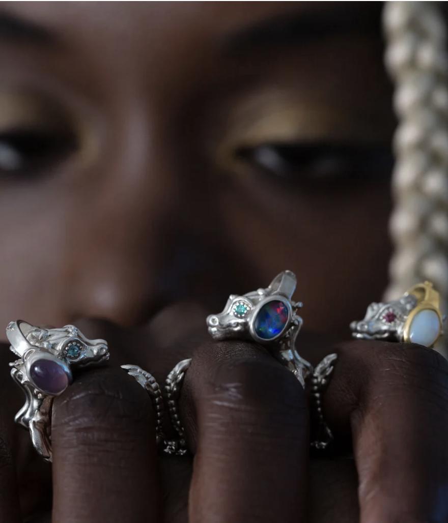 Close-up of a model wearing six sculptural dragon rings in silver and gold finishes, set with opals, turquoise and coloured gemsto