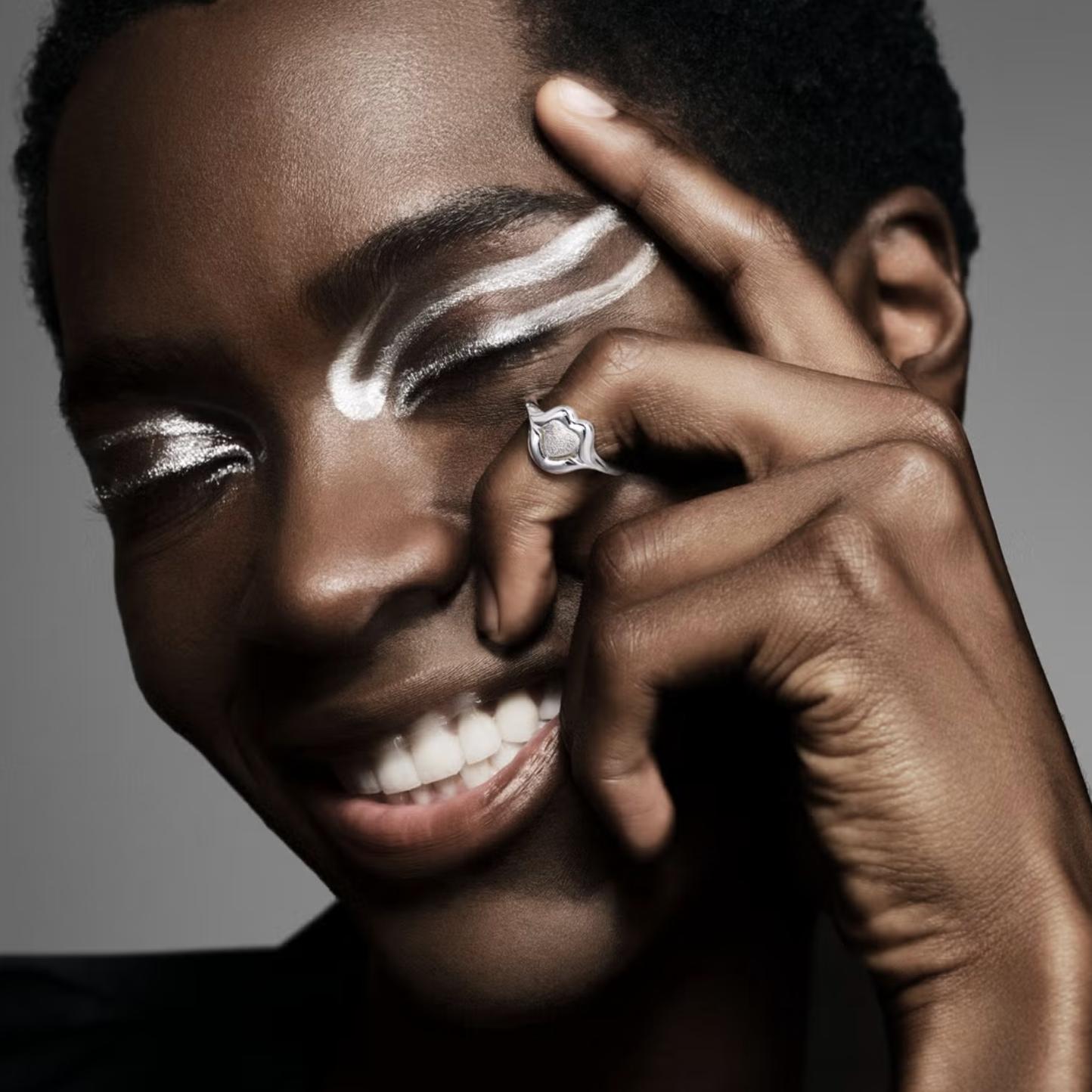 Smiling model with graphic silver eye make-up wearing a sculptural lips-shaped ring on one finger