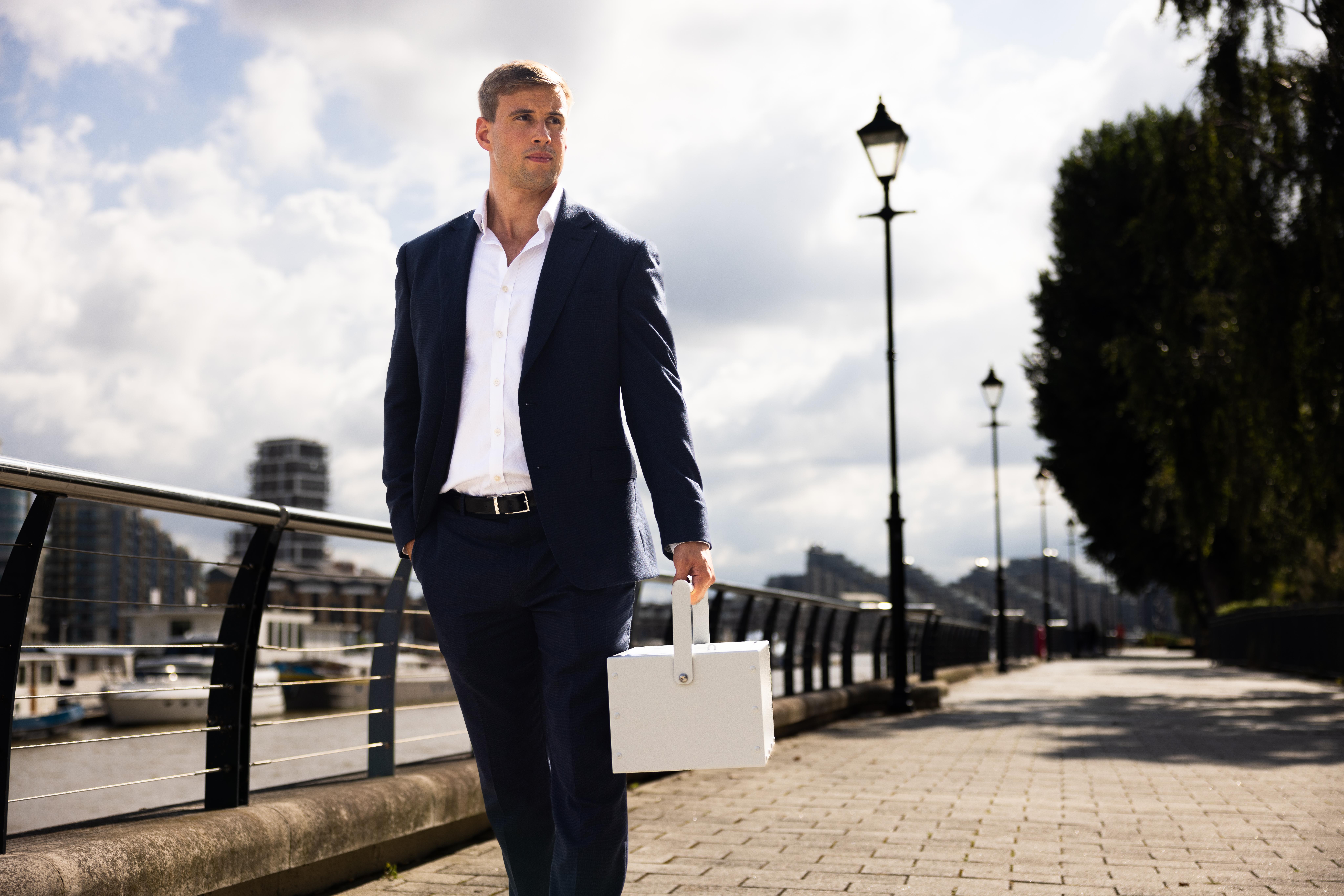 Will Brown of UltraVault, dressed in a navy suit, walking beside the River Thames while carrying a white Portable Safe Deposit Box
