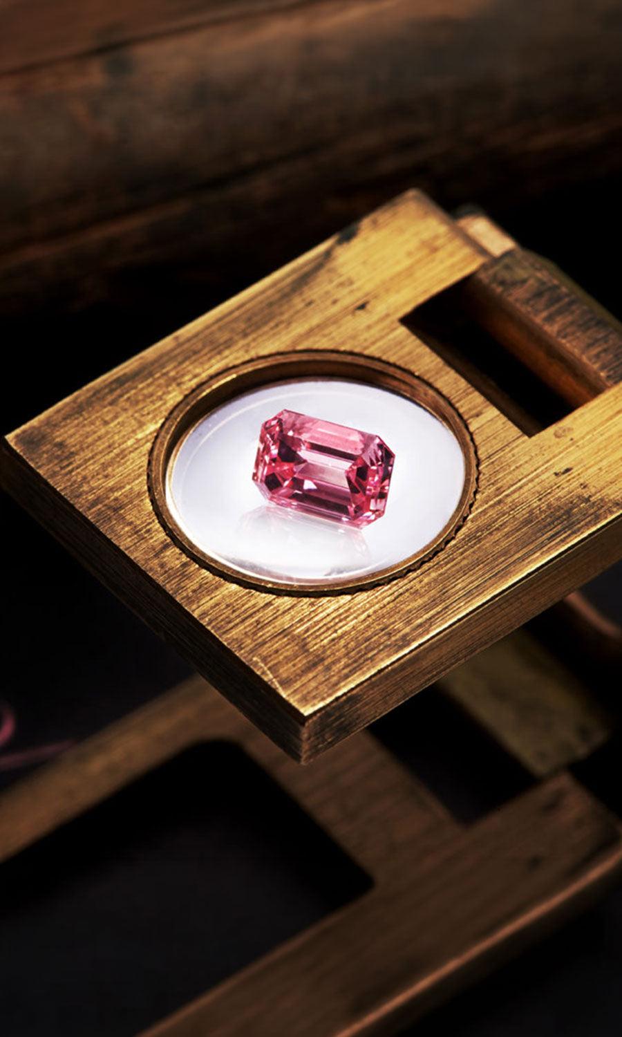 Rectangular step-cut pink diamond resting on a circular white viewing surface within a wooden jeweller’s tool.