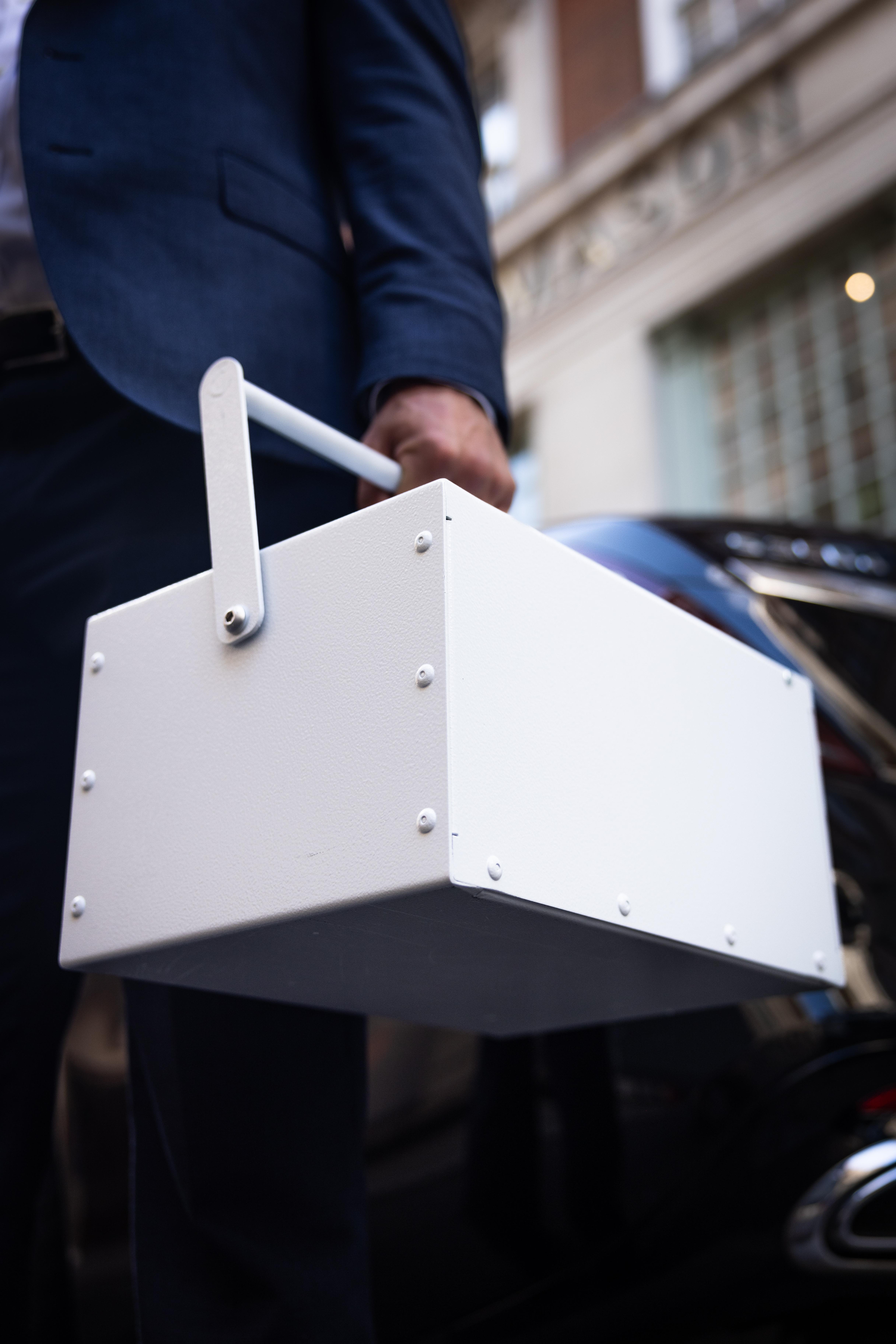 Close-up of a man in a navy suit carrying a white UltraVault Portable Safe Deposit Box beside a car