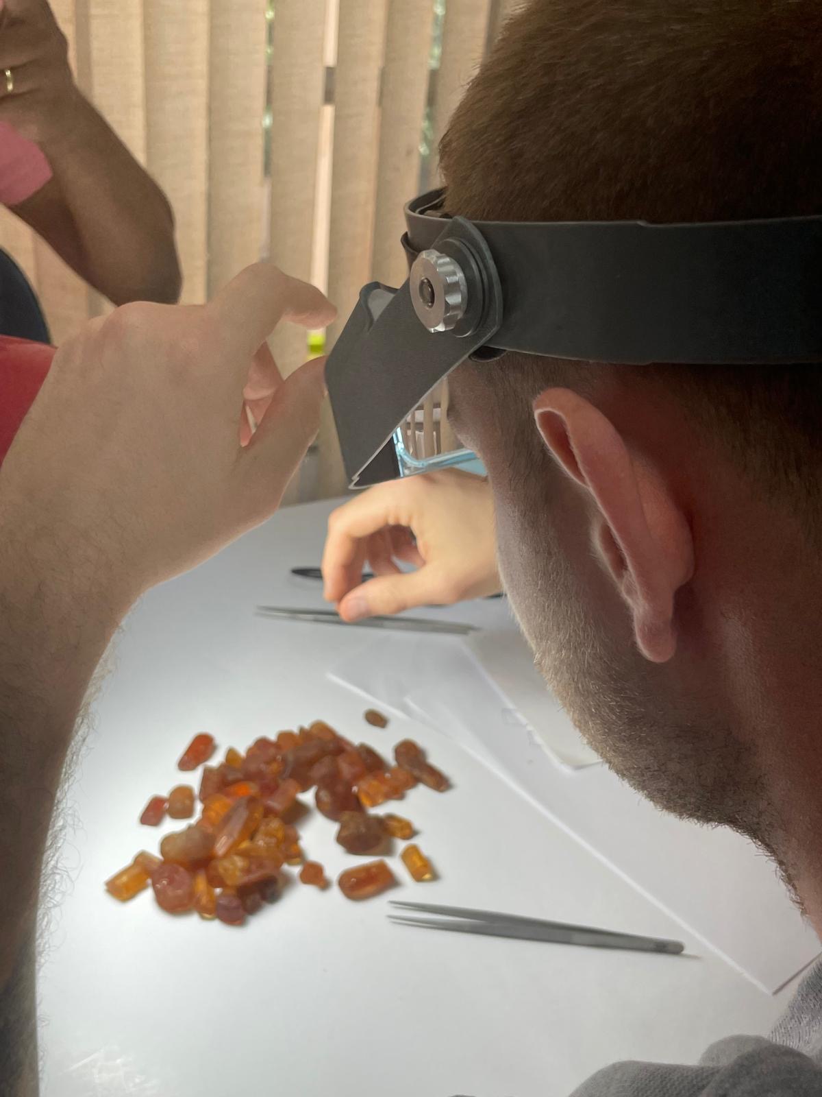 Person examining a pile of rough orange gemstones under magnification, likely spessartine garnets, with tweezers on a white surface.