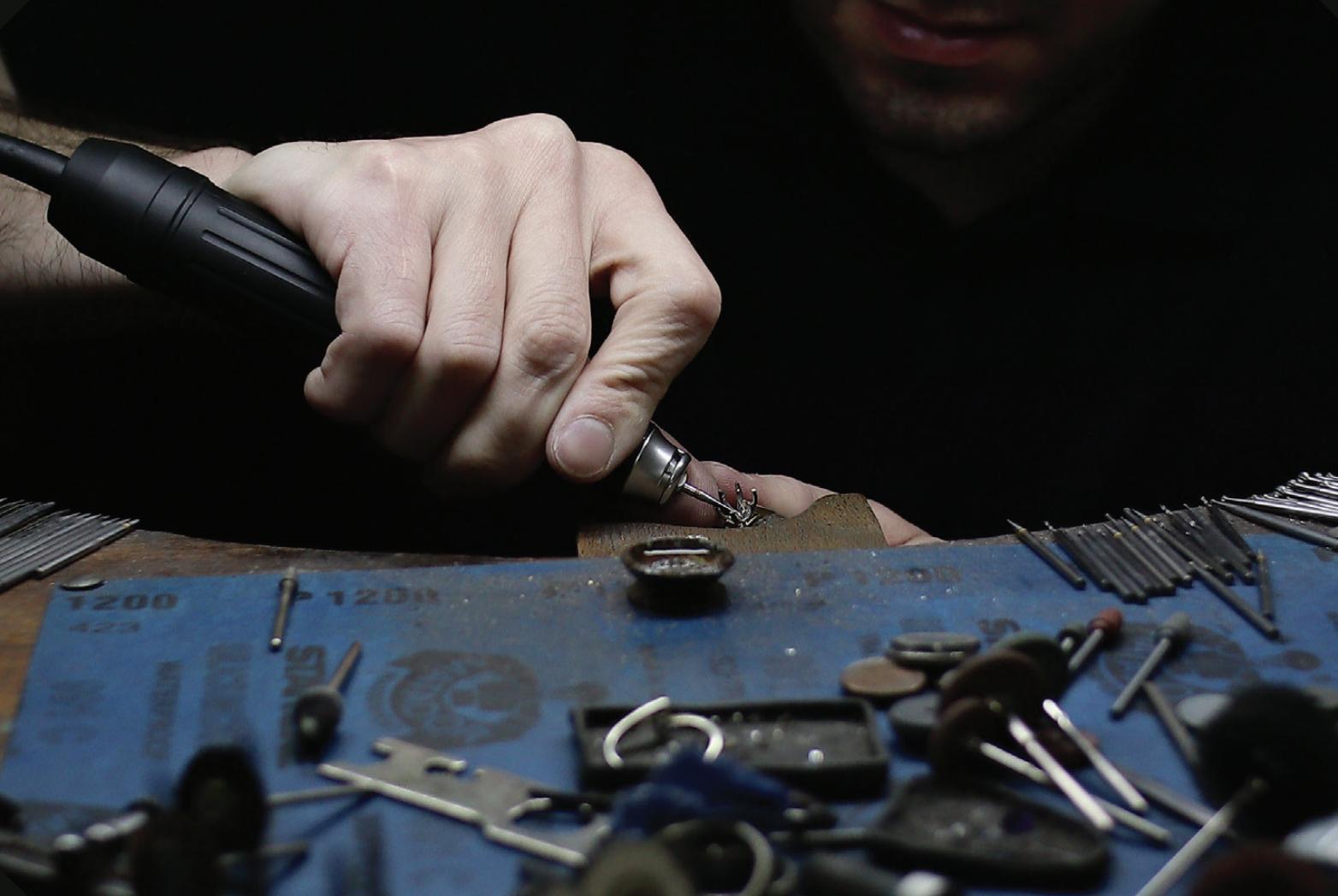 Close-up of a jewellery craftsperson using a rotary tool on a small metal setting at a workbench surrounded by hand tools