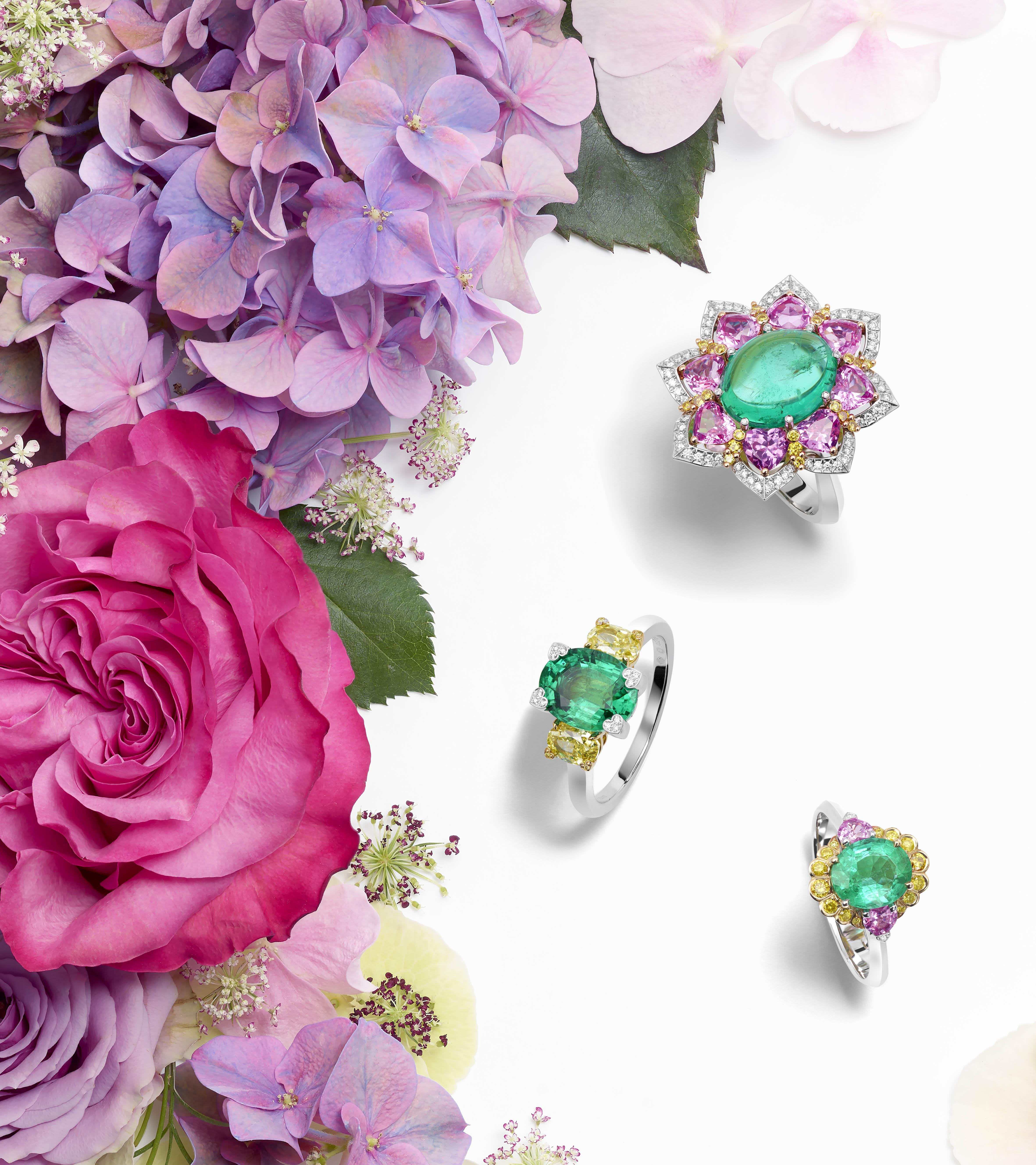 Three emerald rings with pink and yellow sapphires and diamonds arranged on a background of colourful flowers