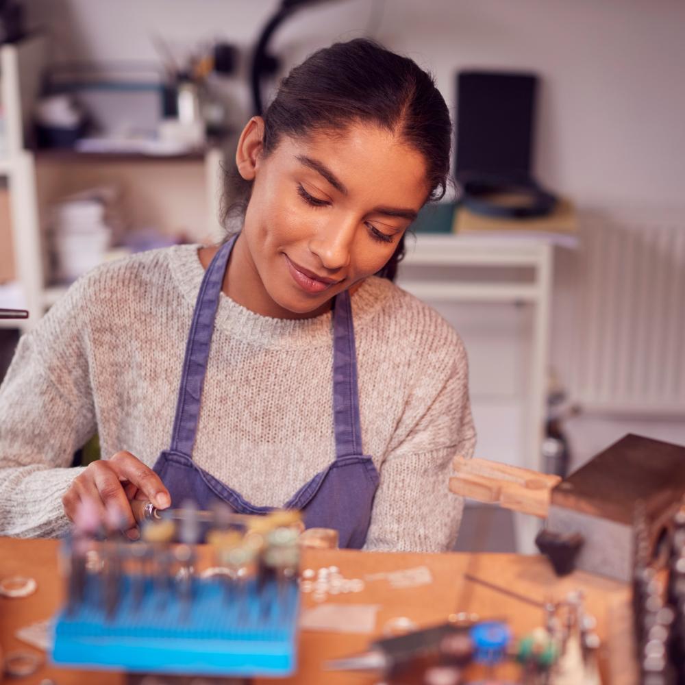 Young jeweller wearing an apron concentrates on detailed work at a bench surrounded by jewellery-making tools