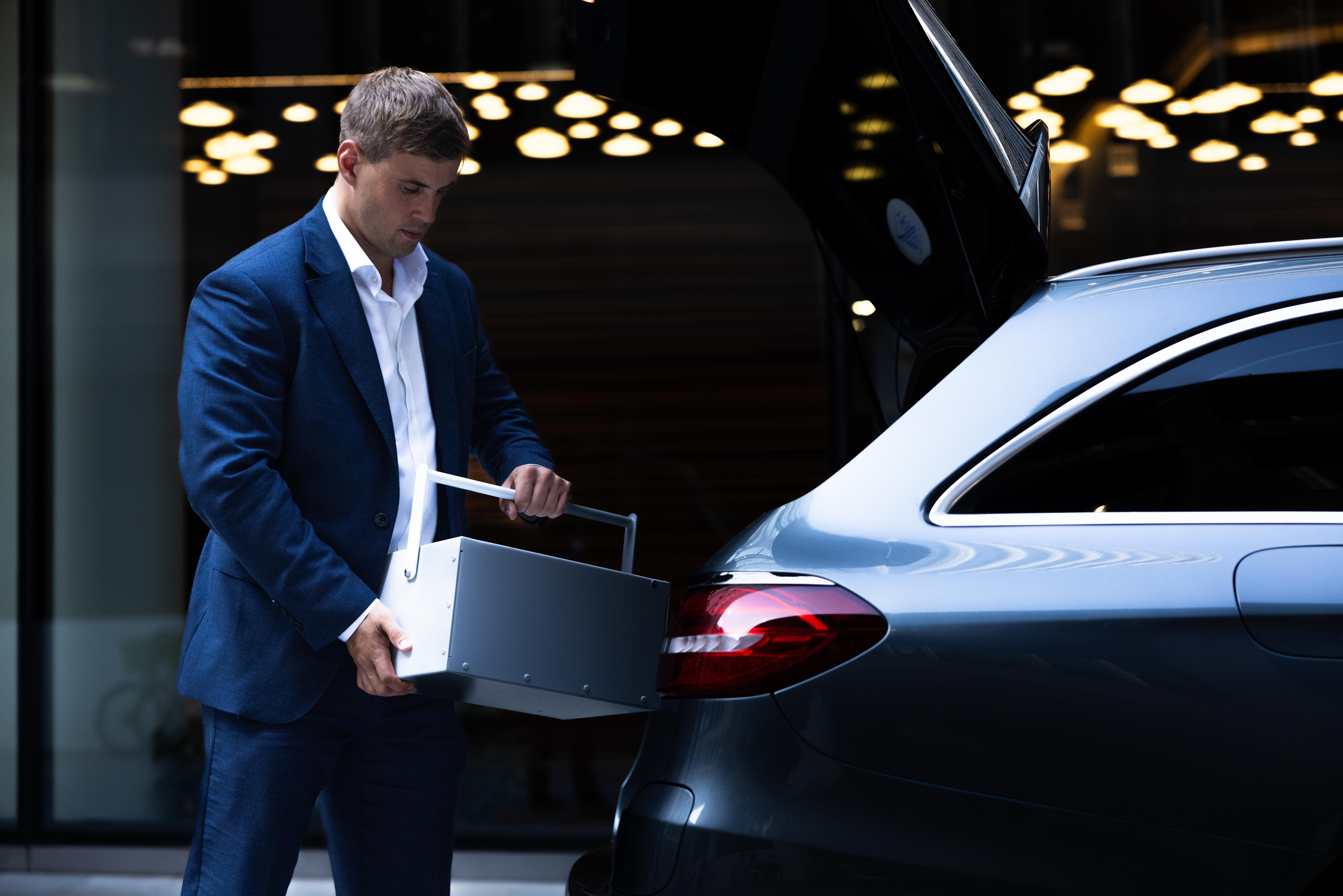 Will Brown, dressed in a navy suit, placing a white UltraVault Portable Safe Deposit Box into the open boot of a car