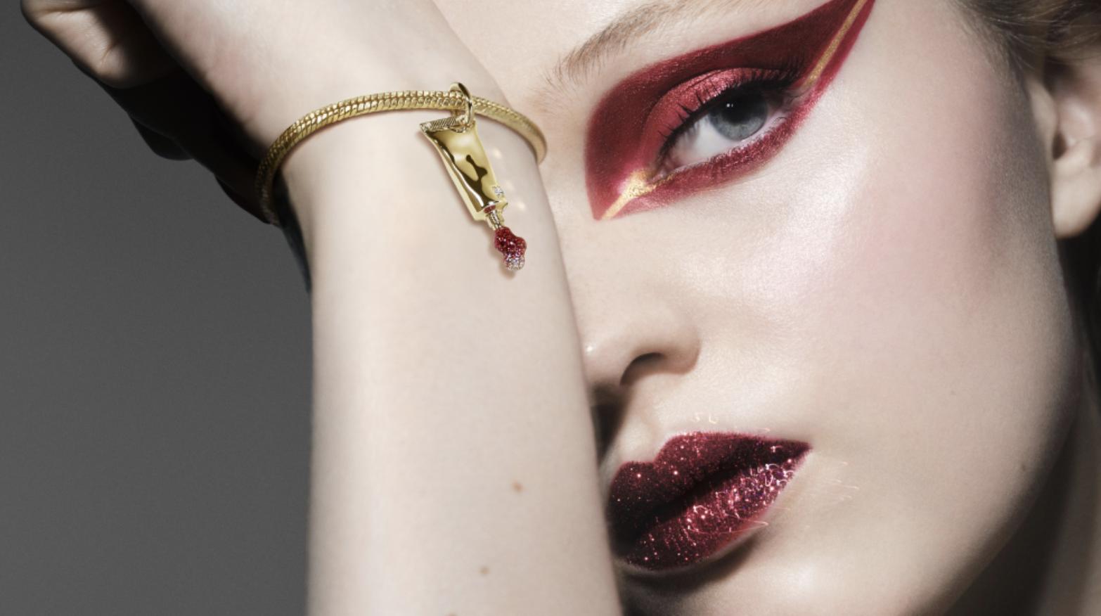 Model wearing dramatic red and gold eye make-up and glittering red lipstick beside a gold bracelet with a dripping beauty tube charm
