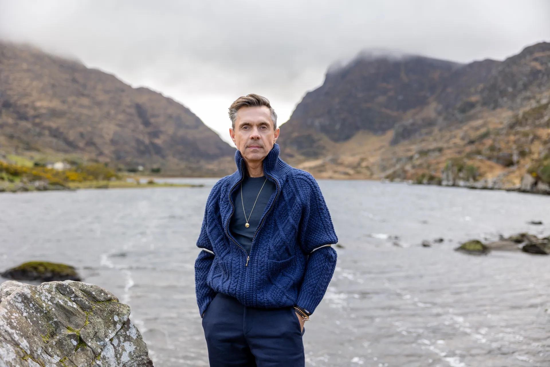 Jewellery designer Shaun Leane wearing a navy knitted jacket beside a mountain lake in rural Ireland.