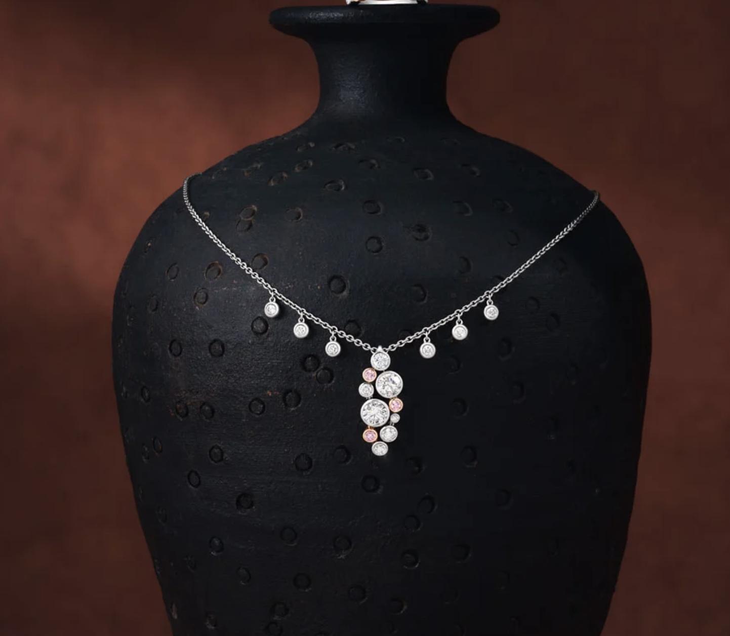 White-diamond drop necklace with small pink diamond accents, displayed on a textured black jewellery bust