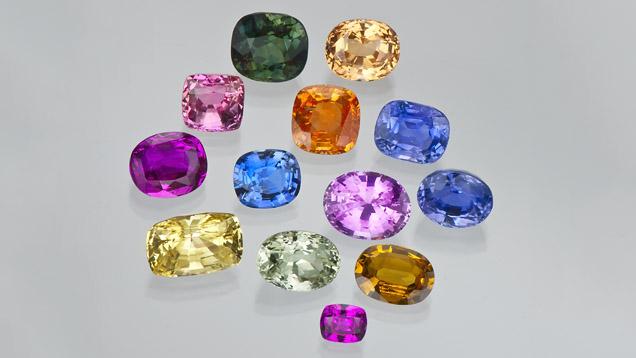 Thirteen faceted sapphires in green, yellow, orange, pink, purple and blue arranged on a pale grey background