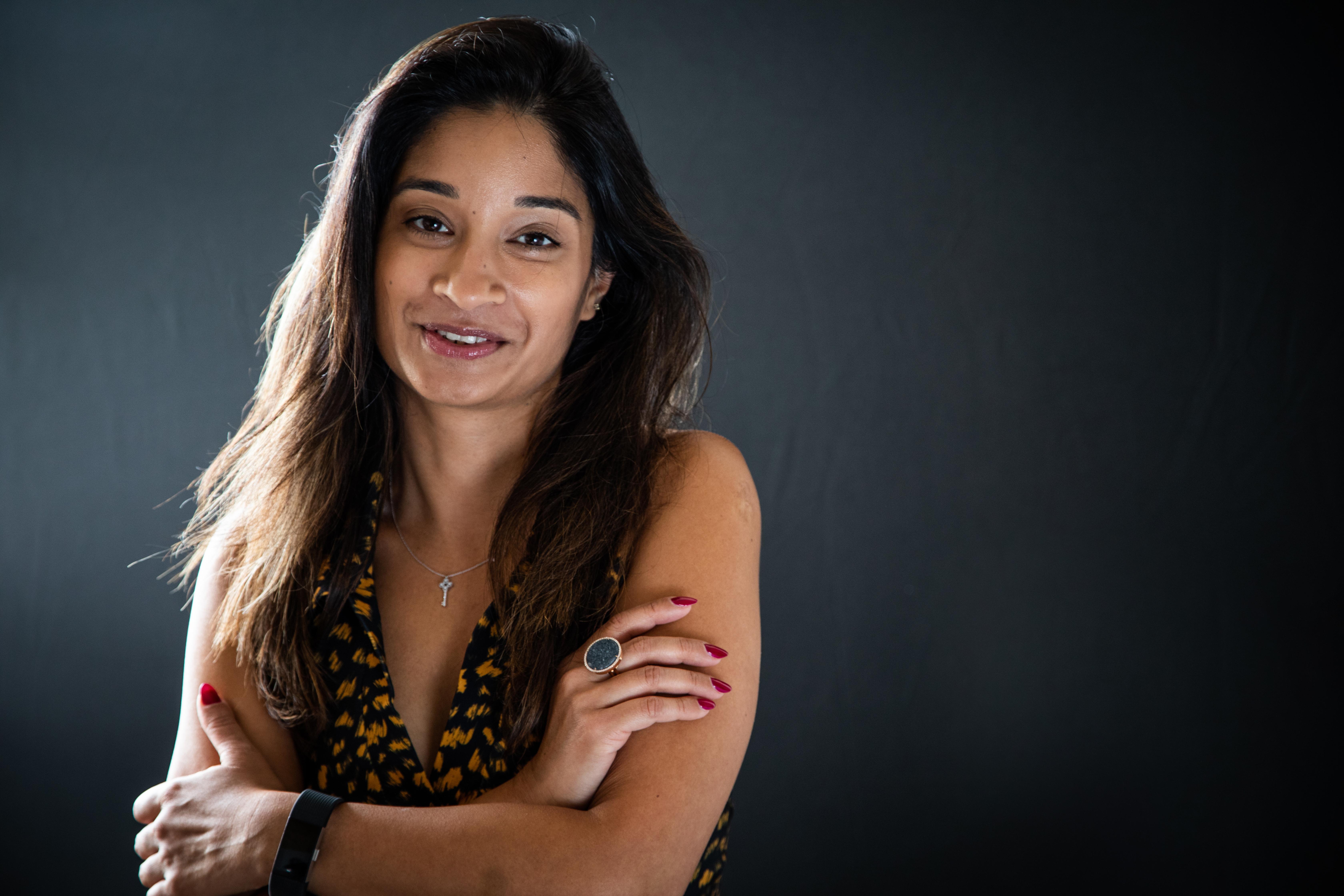 Portrait of Pooja smiling with her arms folded, wearing a black and yellow patterned sleeveless top against a dark grey background