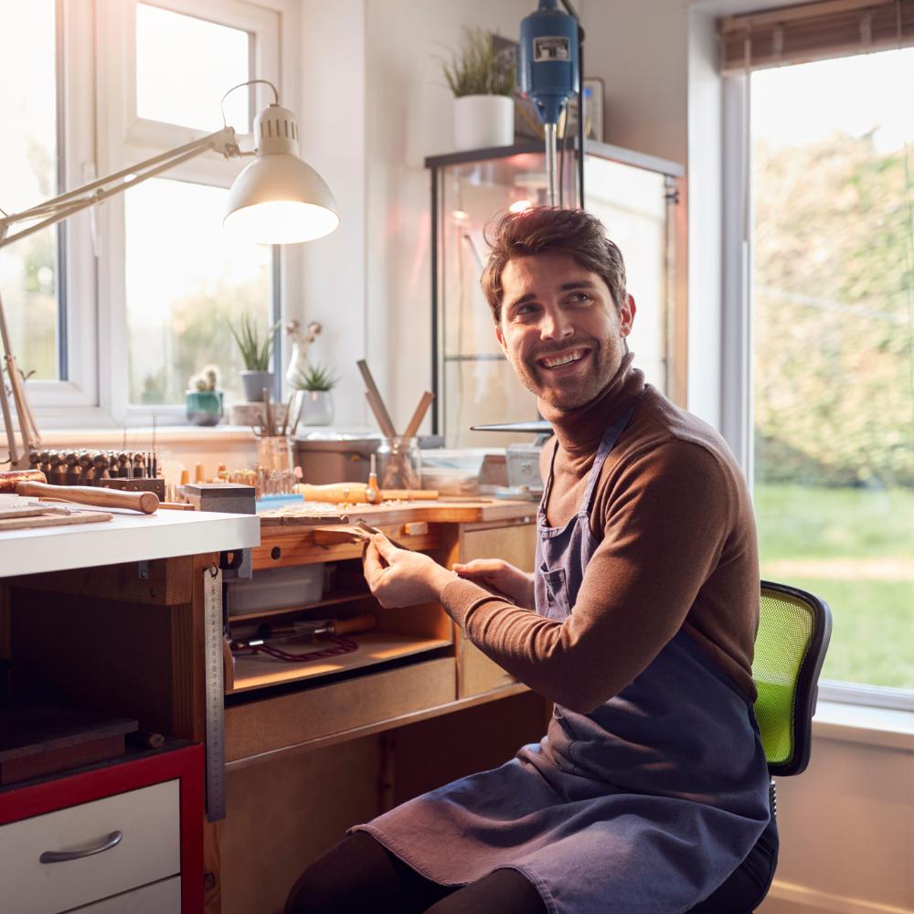 Smiling jeweller wearing an apron and seated at a well-equipped workbench in a bright workshop