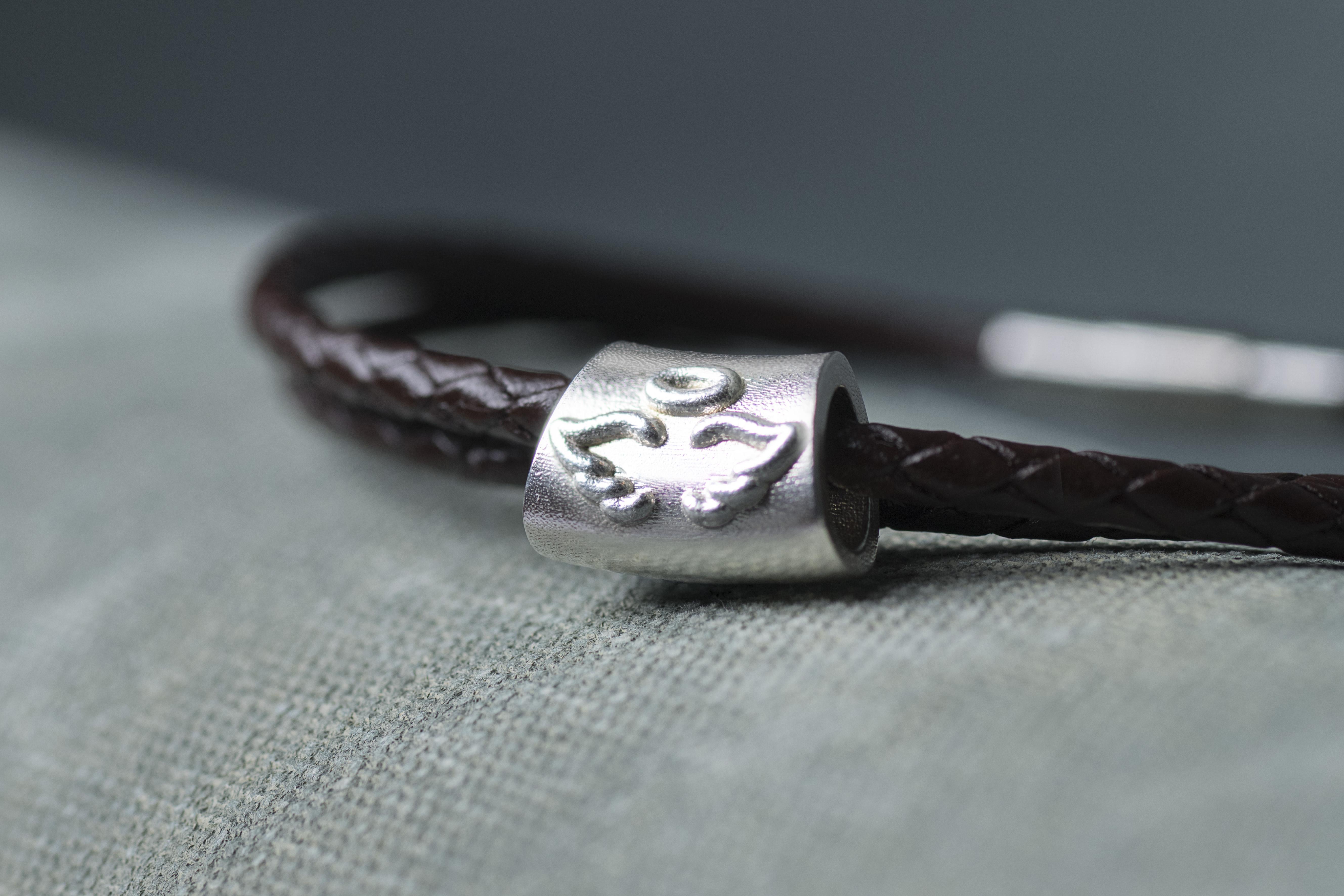 Close-up of the silver Angel Baby Charm with raised angel wings and a halo, threaded onto a brown braided leather bracelet on a pale grey surface.