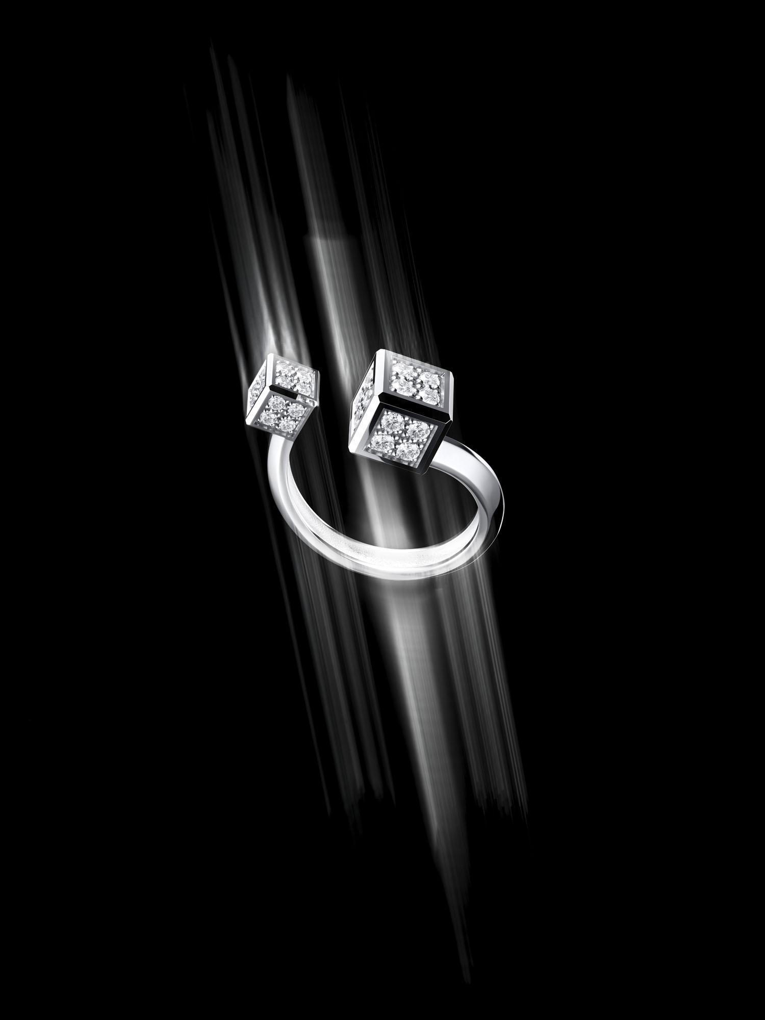 An open ring in white metal featuring two diamond-set cube ends, set against a black background with light streaks highlighting its curved form.