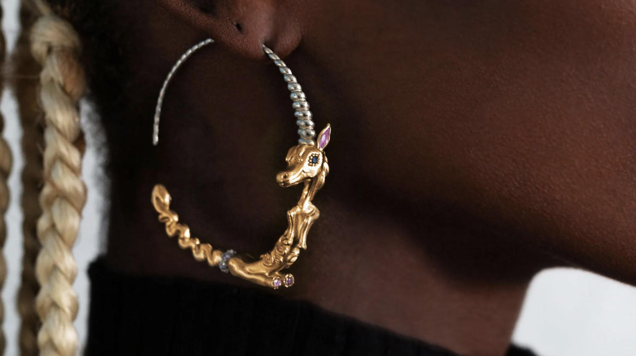 Close-up of a large hoop earring shaped as a gold unicorn with a twisted silver horn and colourful gemstone details.