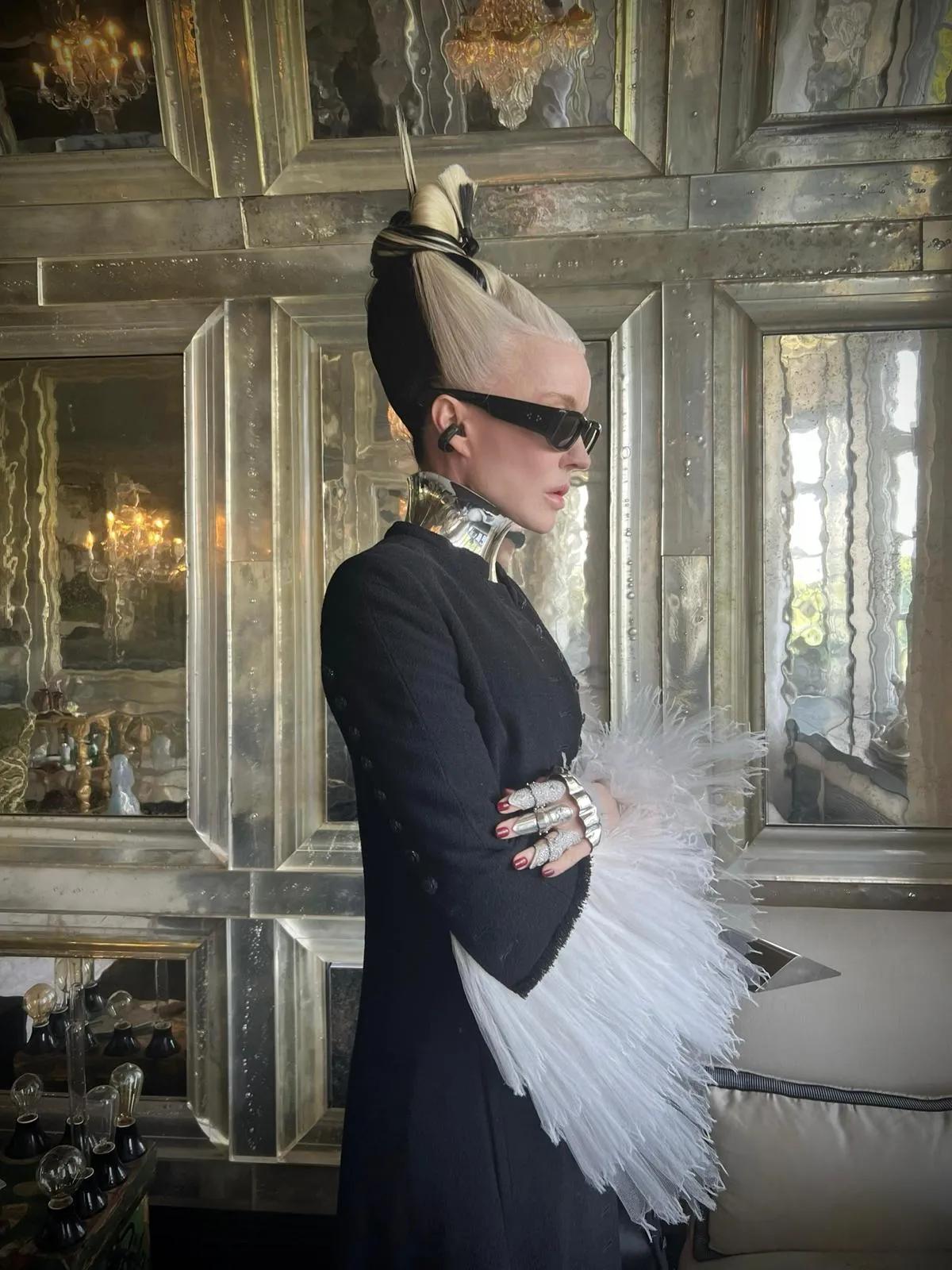 Daphne Guinness in profile wearing dark sunglasses, a black outfit and a sculptural silver collar, standing in a mirrored interior