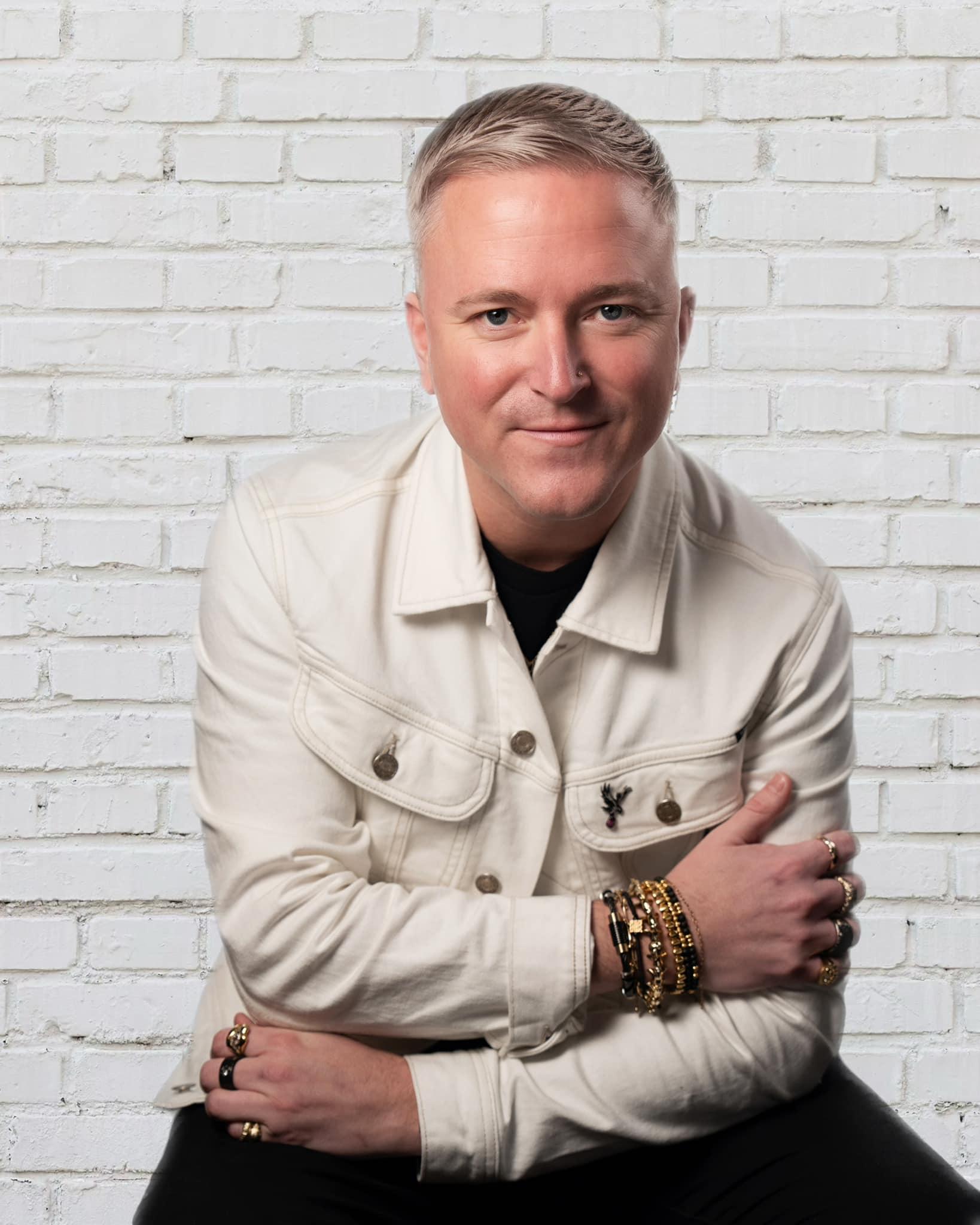 Portrait of Andrew Martyniuk wearing a cream jacket and gold jewellery, seated against a white brick wall