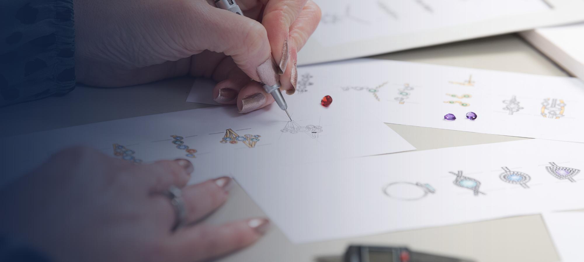 A Weston Beamor designer sketches a bespoke jewellery concept by hand beside a loose red gemstone and coloured design drawings.