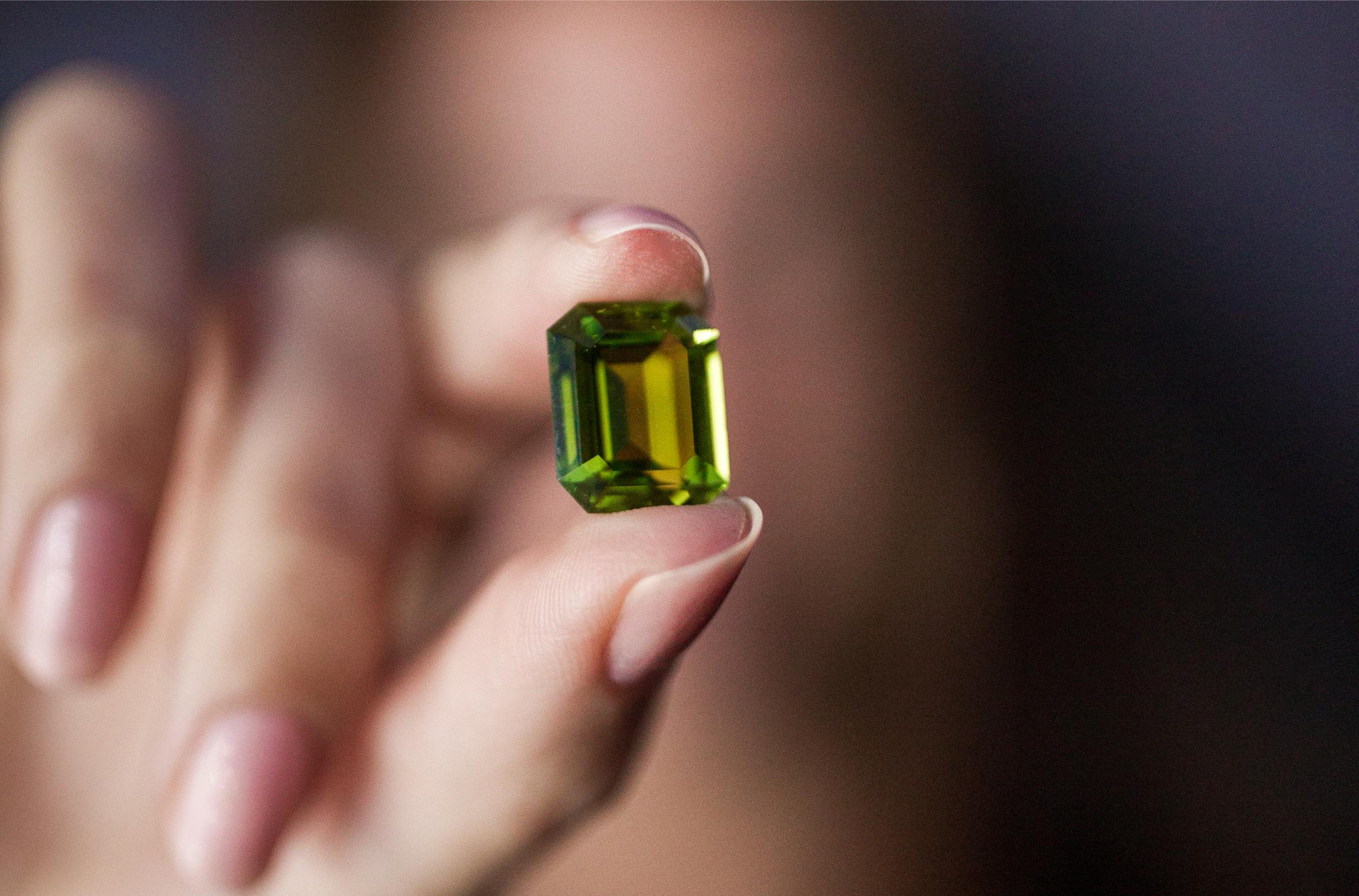 Close-up of a rectangular emerald-cut peridot held between two fingers, showing deep olive-green and golden-green tones against a softly blurred background.