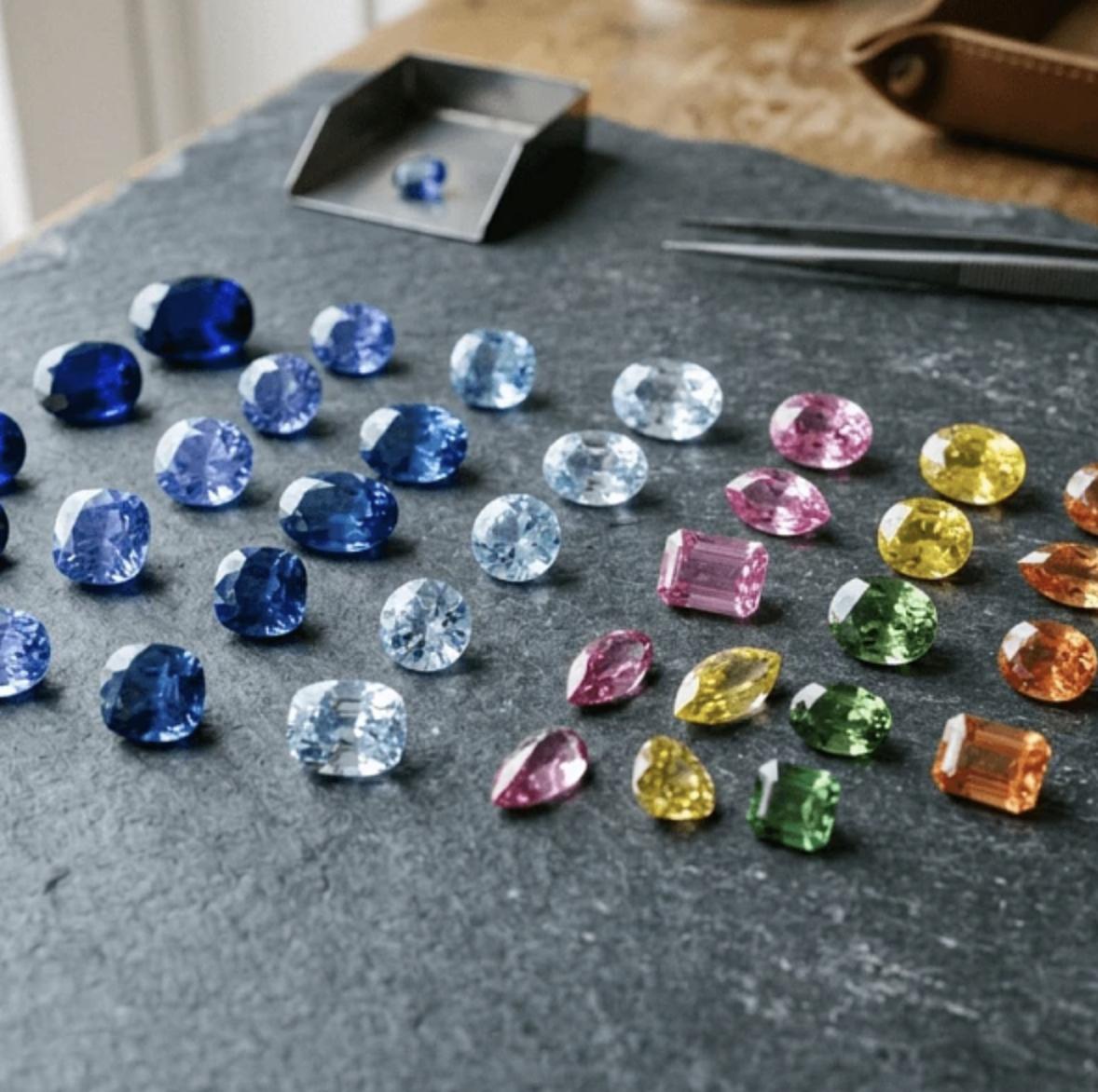 Loose blue, aqua, pink, yellow, green and orange sapphires arranged on a dark jeweller&rsquo;s tray beside tweezers.