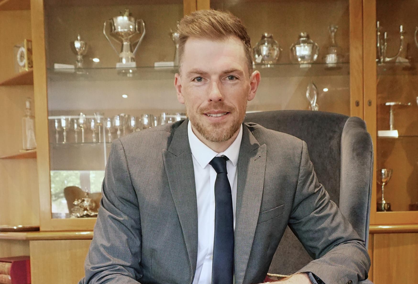 Portrait of NAJ Chief Executive Ben Massey seated in a grey suit and dark tie, with a cabinet of trophies behind him