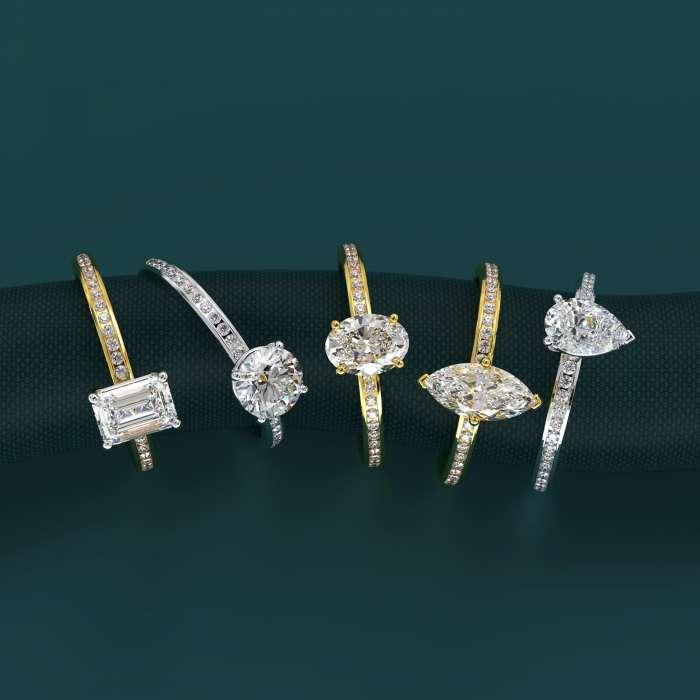 Five finished diamond engagement rings in yellow and white metal, featuring emerald, round, oval, marquise and pear-shaped centre stones.